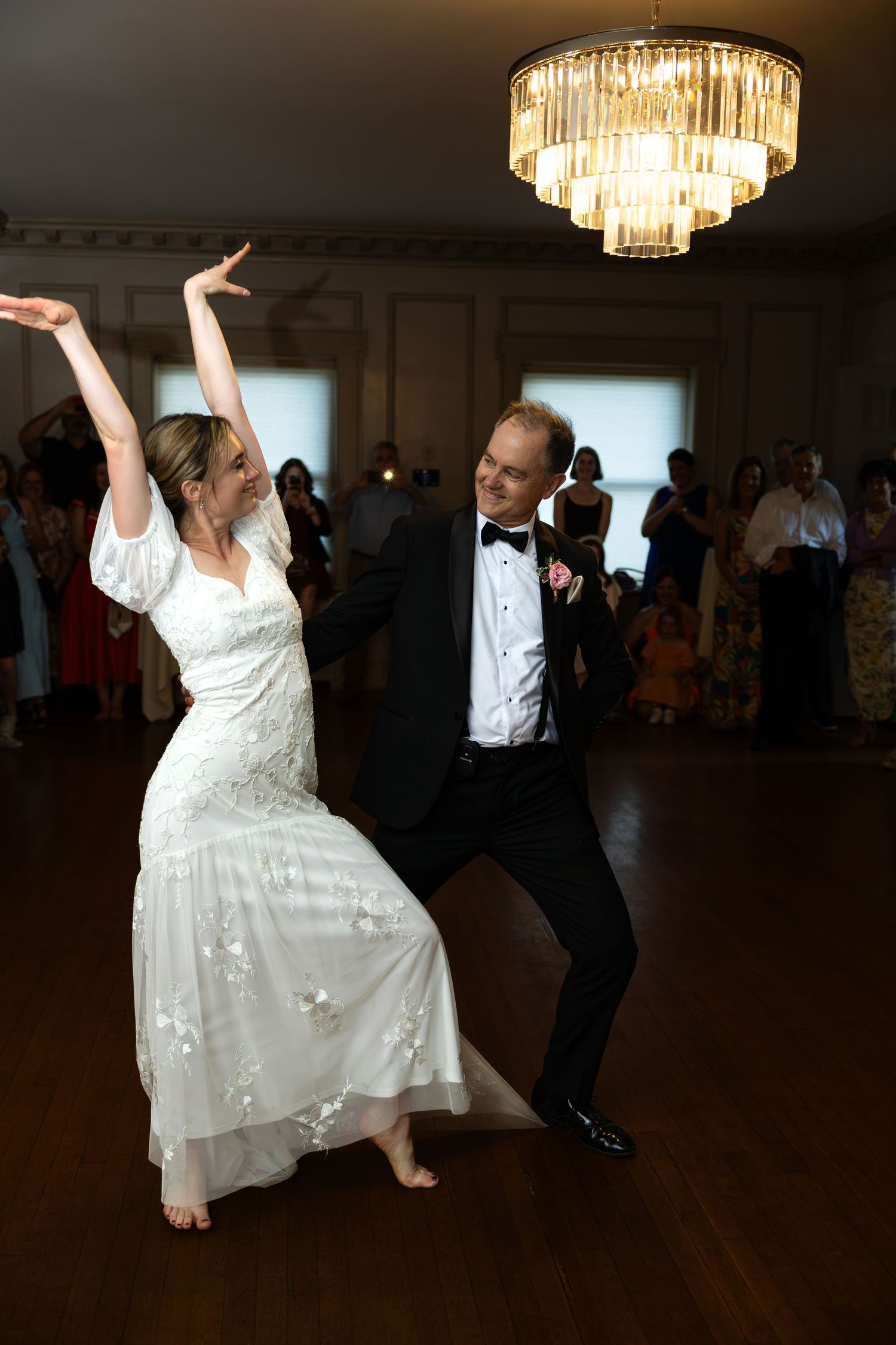 A couple performs an energetic dance move under an elegant chandelier at their reception.