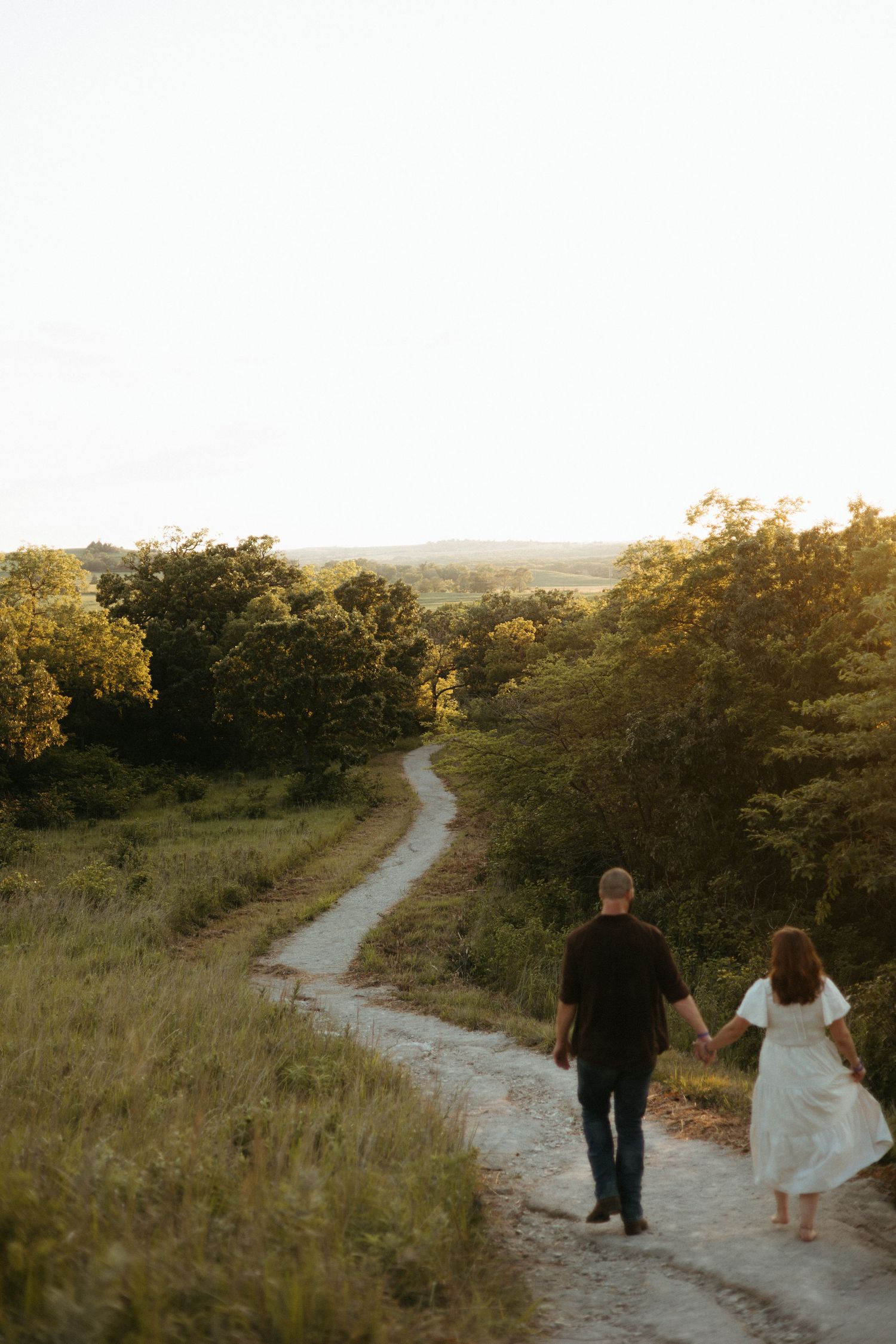 A couple walks hand in hand down a winding dirt path through golden hills at sunset.