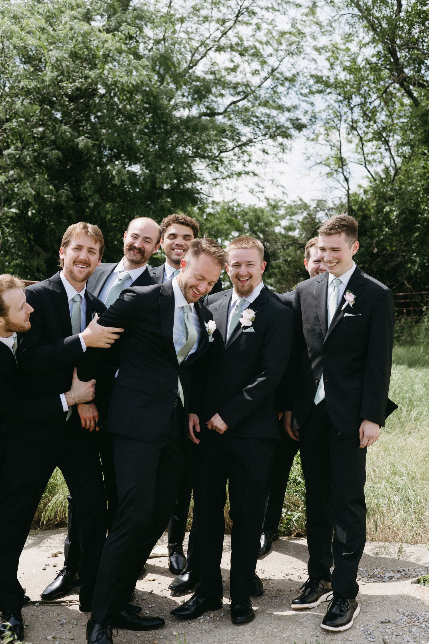 Groom laughing with groomsmen in dark suits and sage green ties outdoors on a sunny wedding day.