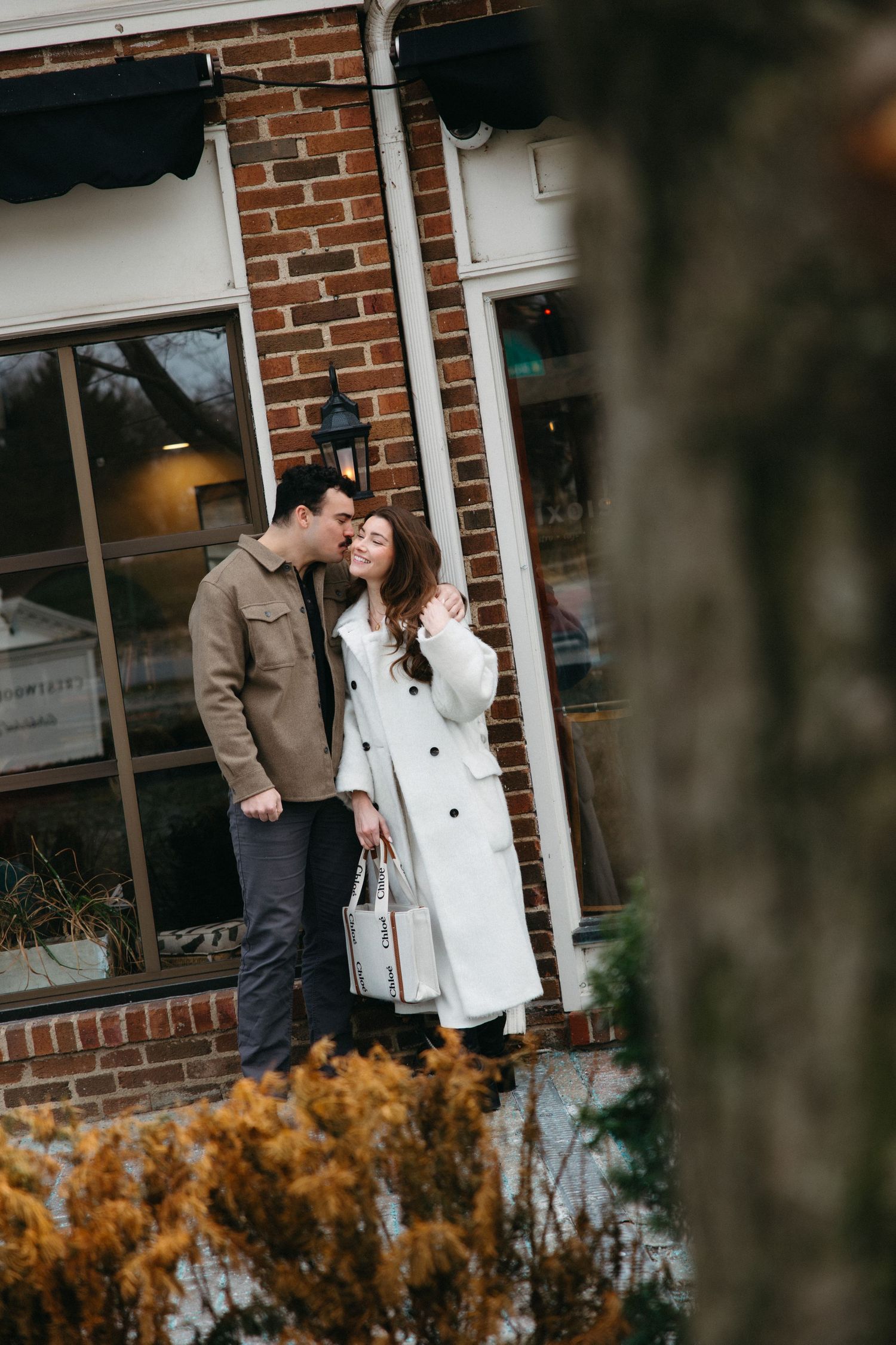 A couple shares a romantic moment outside a brick building with autumn foliage in the foreground.