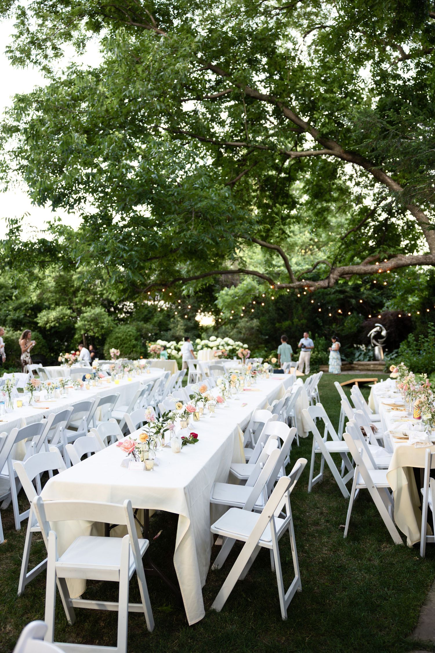 Outdoor wedding reception setup with white chairs and tables under large trees.