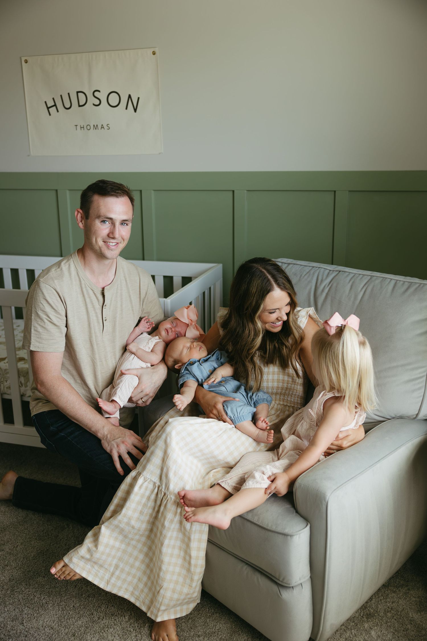 A family relaxes together on a light gray couch in a room with soft green walls and natural lighting.