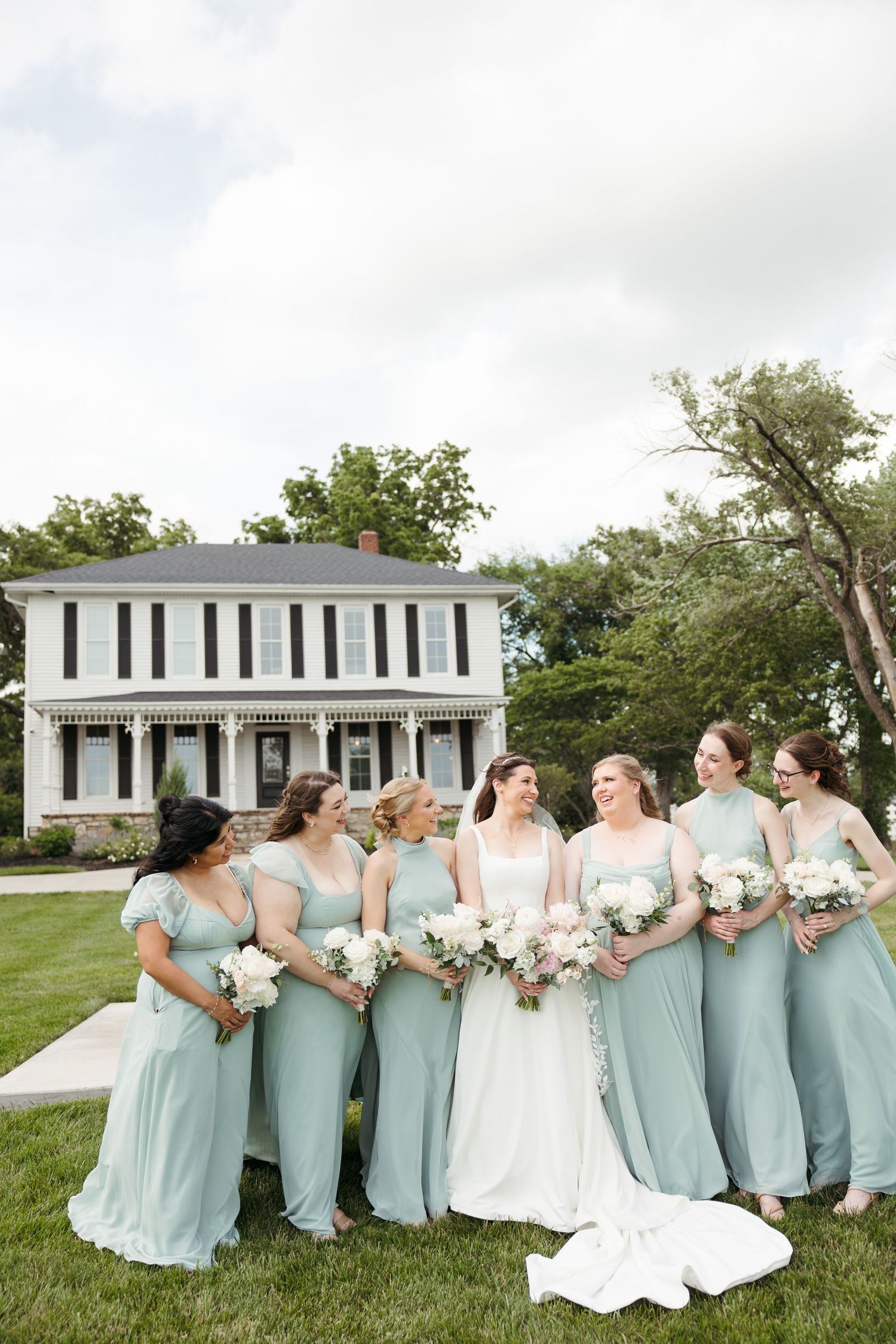 Bride in white gown with bridesmaids in sage green dresses holding white bouquets outside a classic white farmhouse.