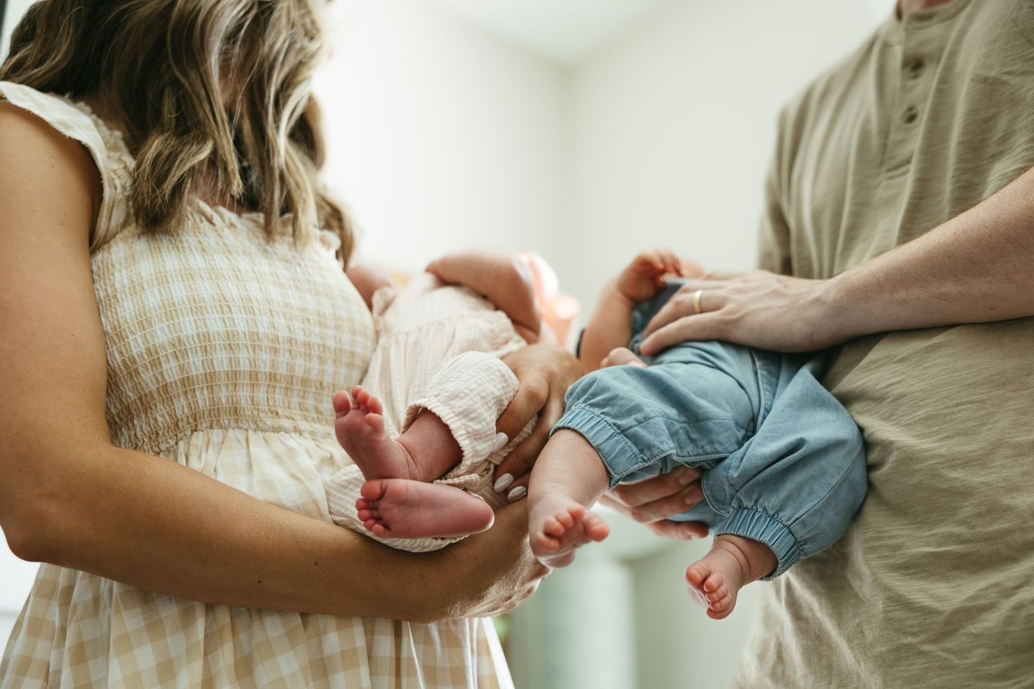 A newborn baby in denim shorts is gently cradled by two pairs of caring hands against a white background.