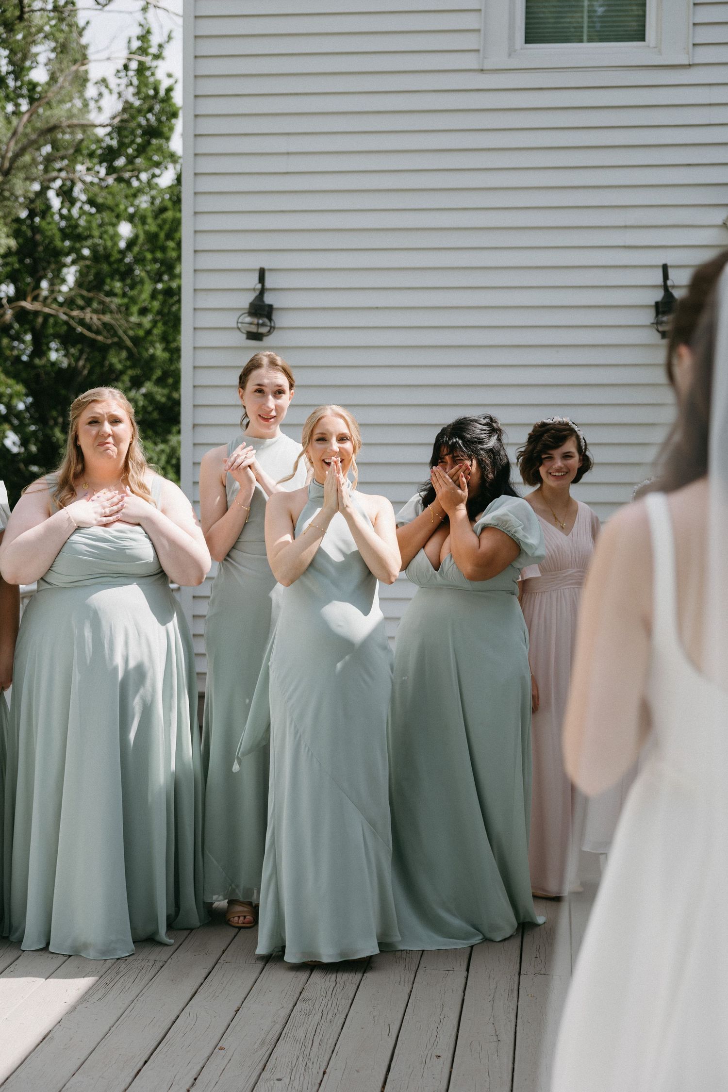 Bridesmaids in sage green gowns react with joy and emotion as they see the bride for the first time on her wedding day.