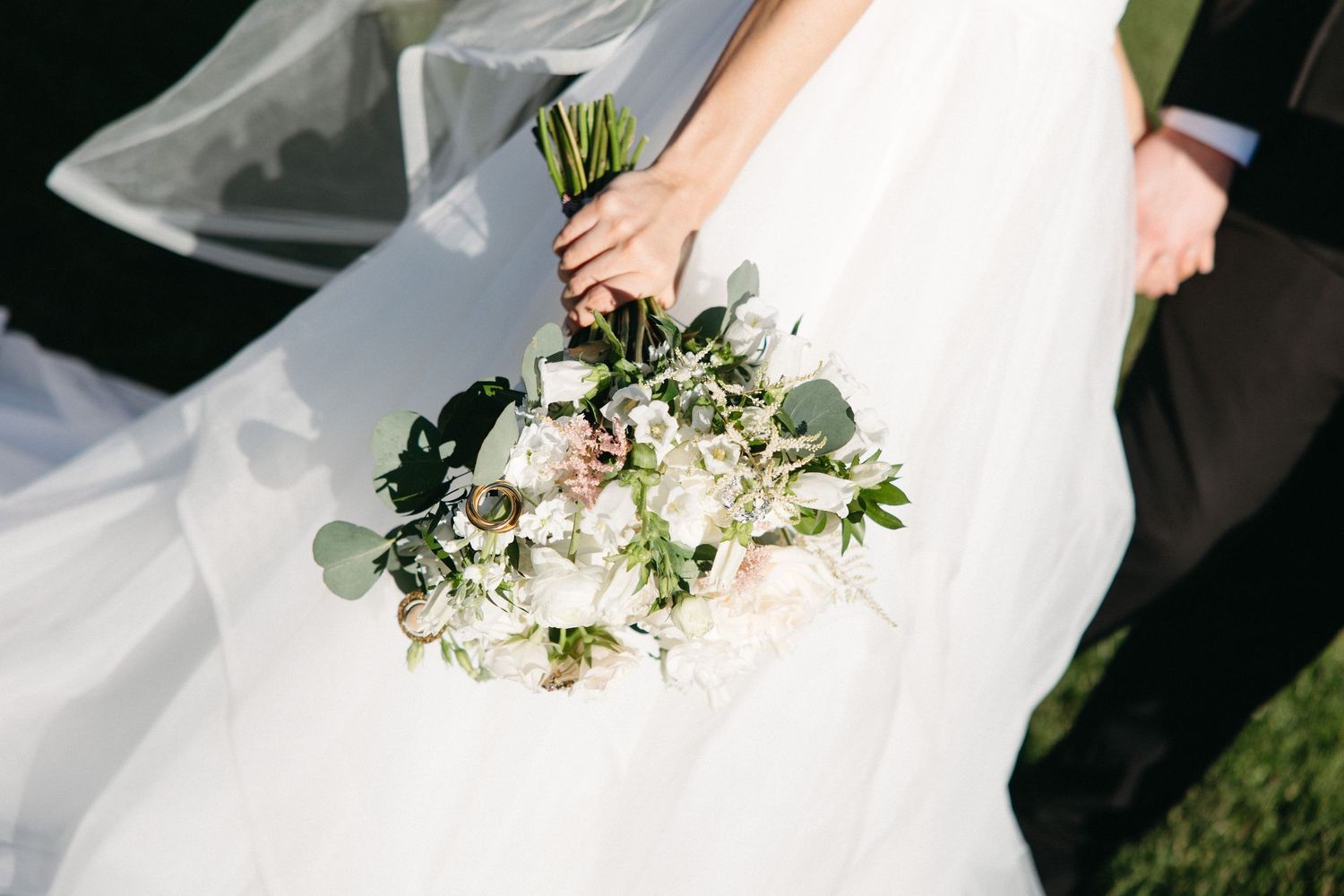A bride in a flowing white dress holds a delicate bouquet of white roses and eucalyptus against a dark background.