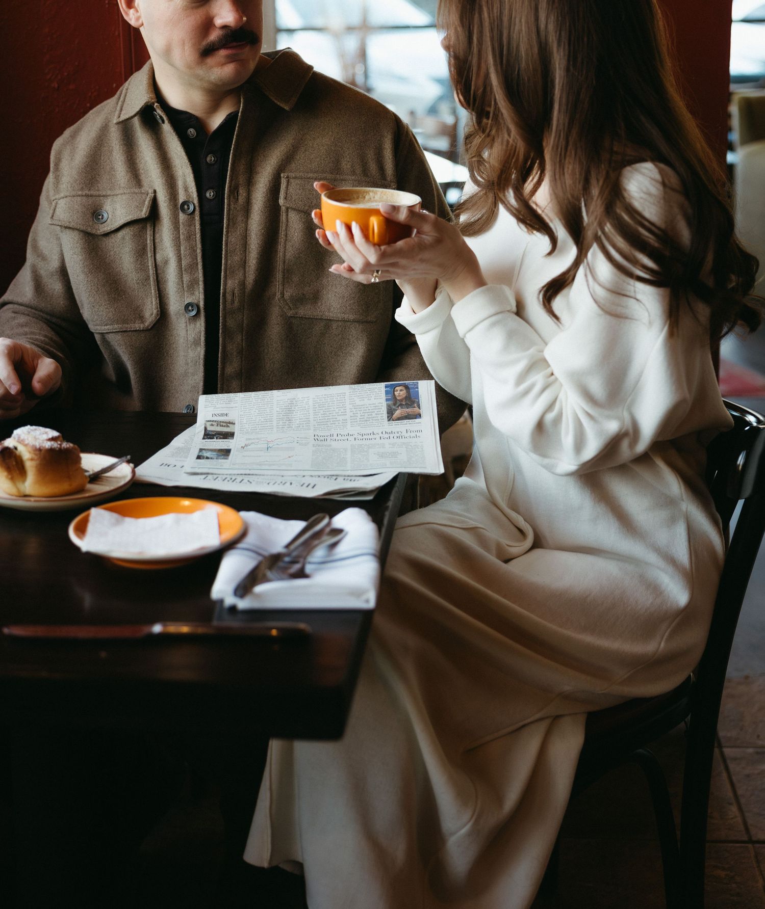 Two people sit at a dark wooden table in a cafe reviewing paperwork while having coffee and pastries.