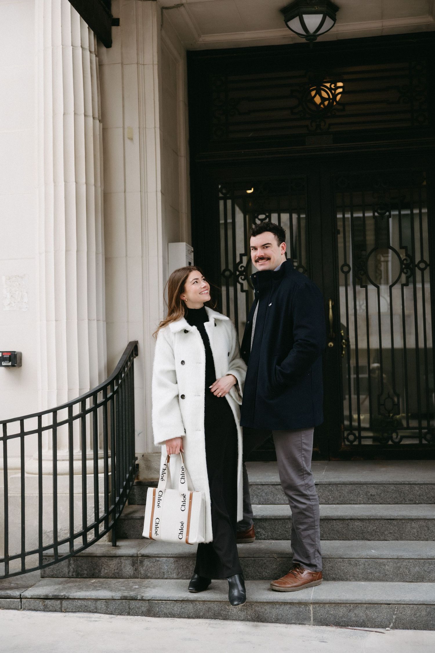 Two people stand on stone steps outside a building entrance with white columns and black railings.