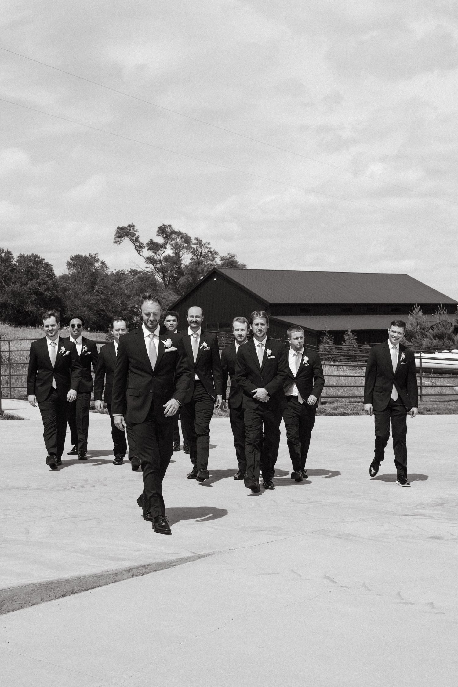 Black and white photo of a groom leading groomsmen in suits walking confidently outdoors near a barn venue.