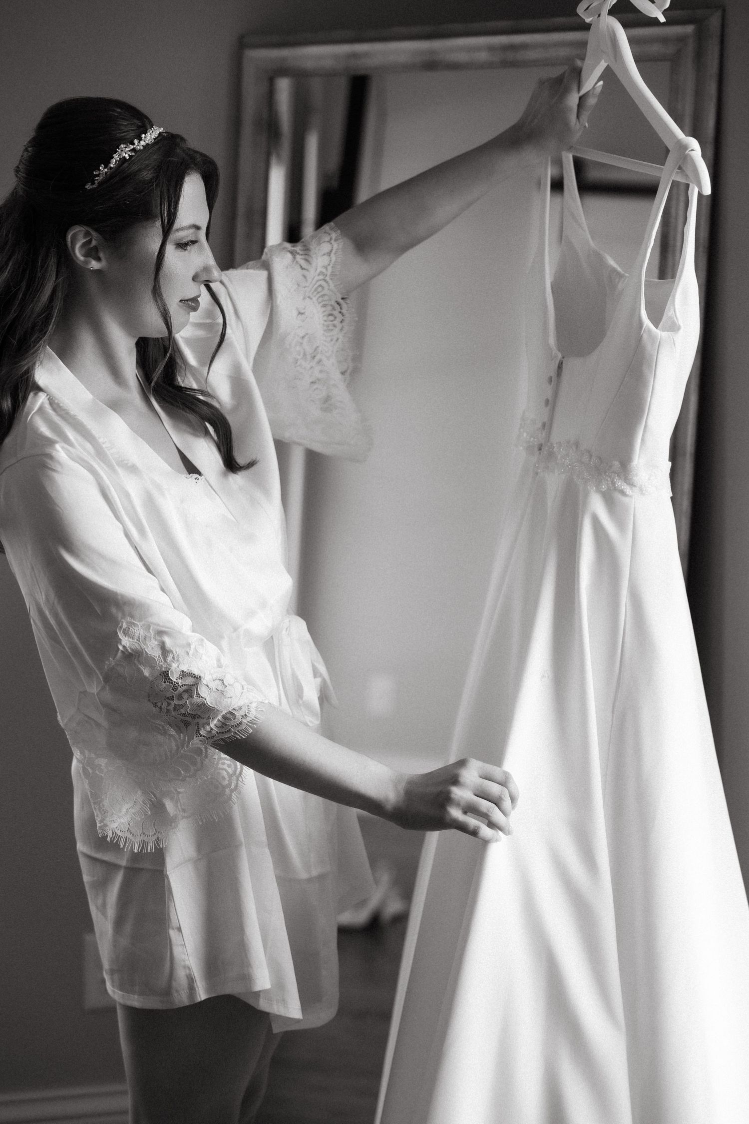 Bride in lace robe admiring her wedding dress on a hanger in black and white photo.
