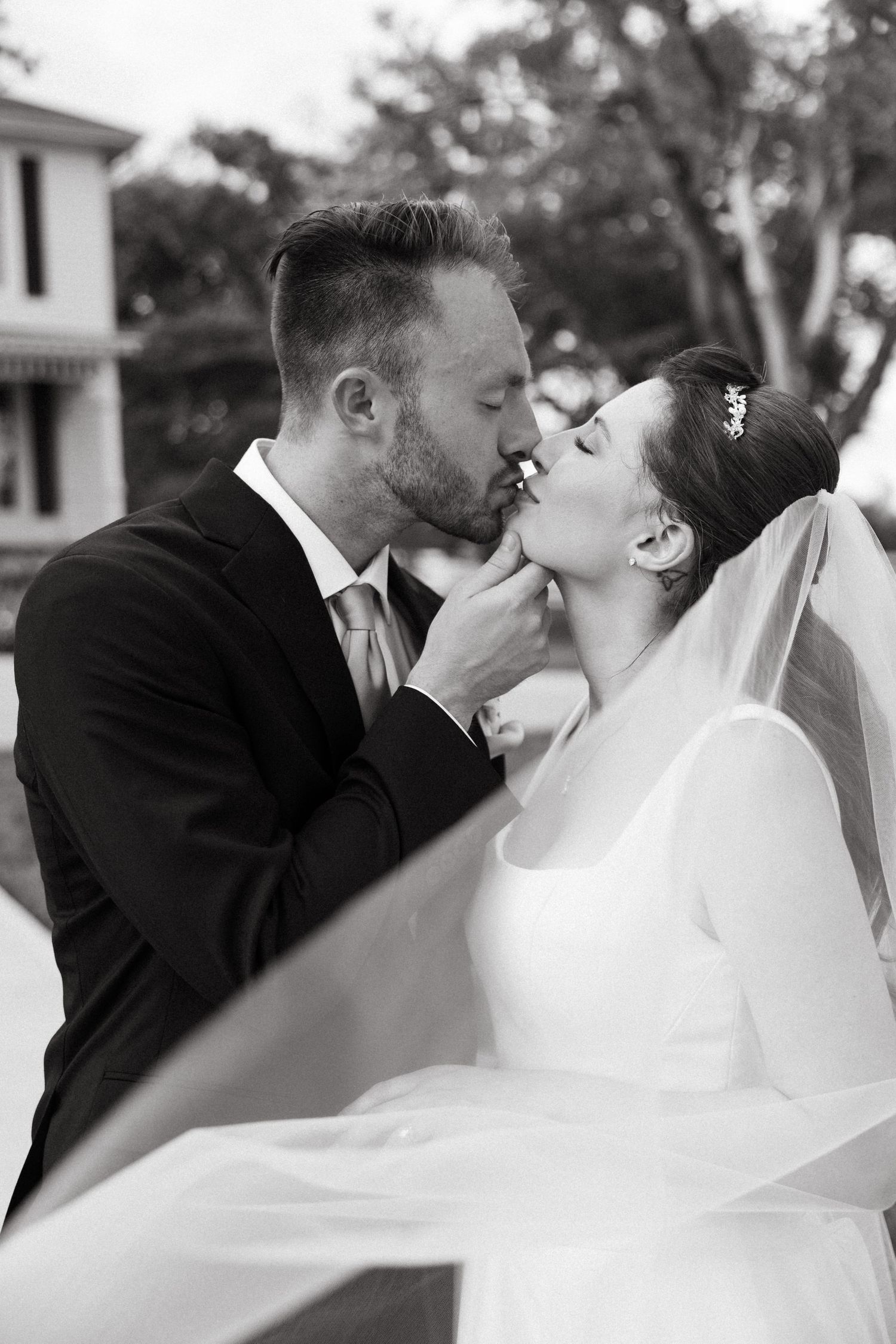 Black and white photo of bride and groom sharing a romantic kiss, with veil flowing in the breeze.