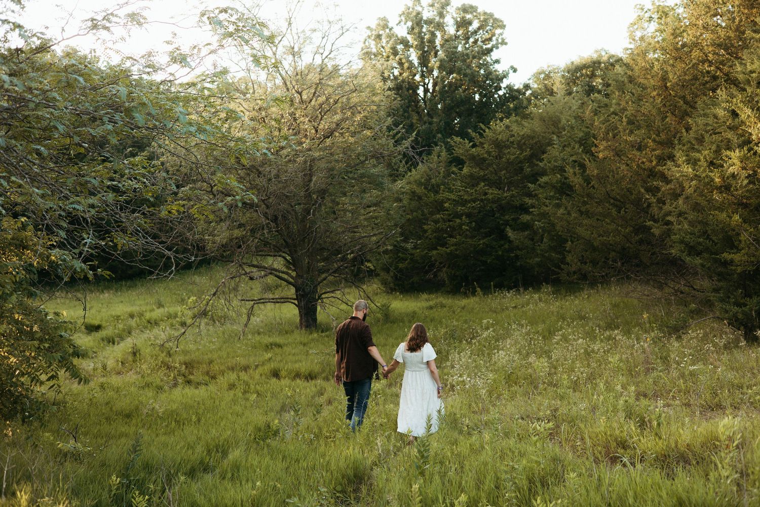 A couple walking through a lush green field surrounded by trees.