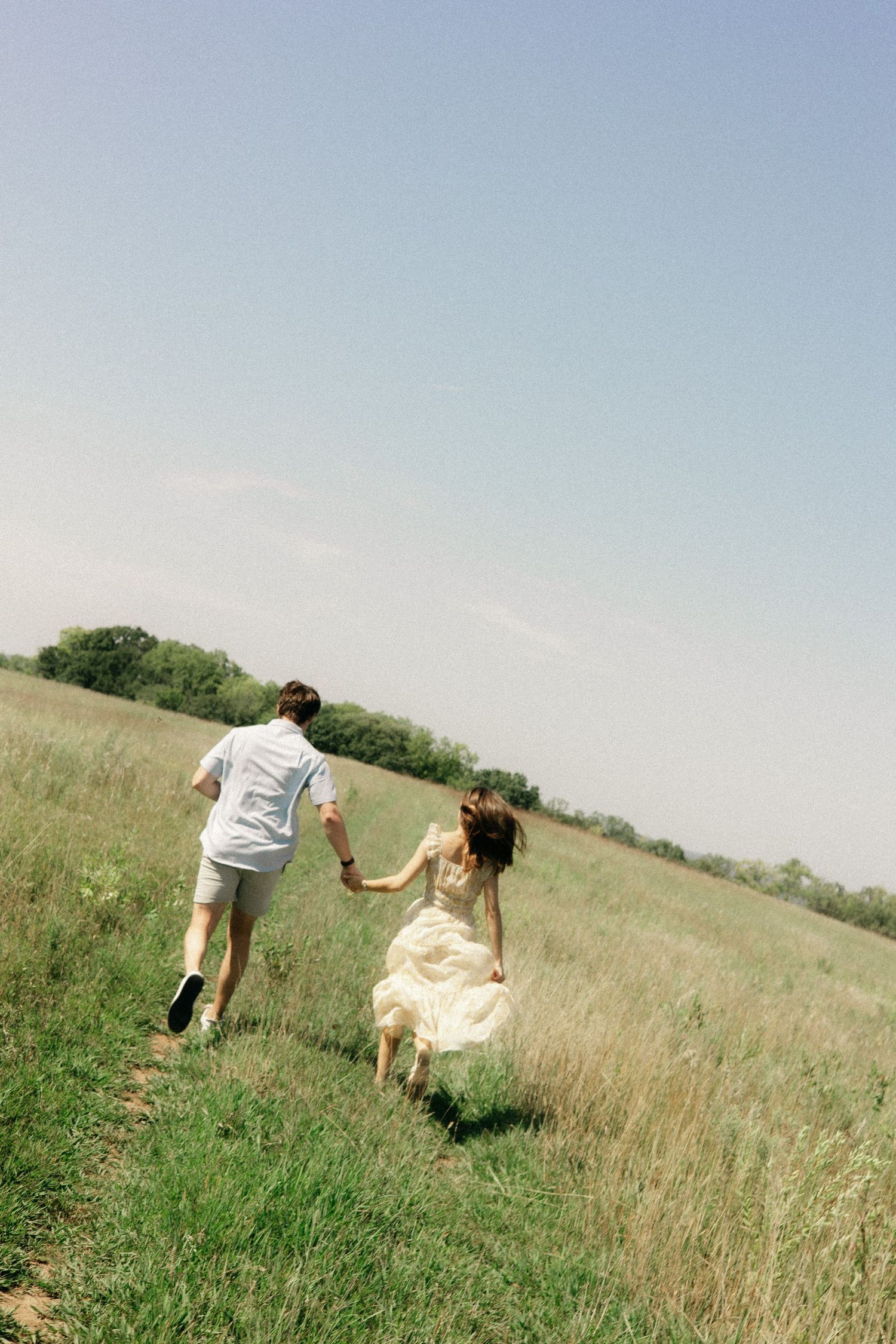 A couple runs hand in hand through a sunny meadow, with the person in white wearing a flowing dress.