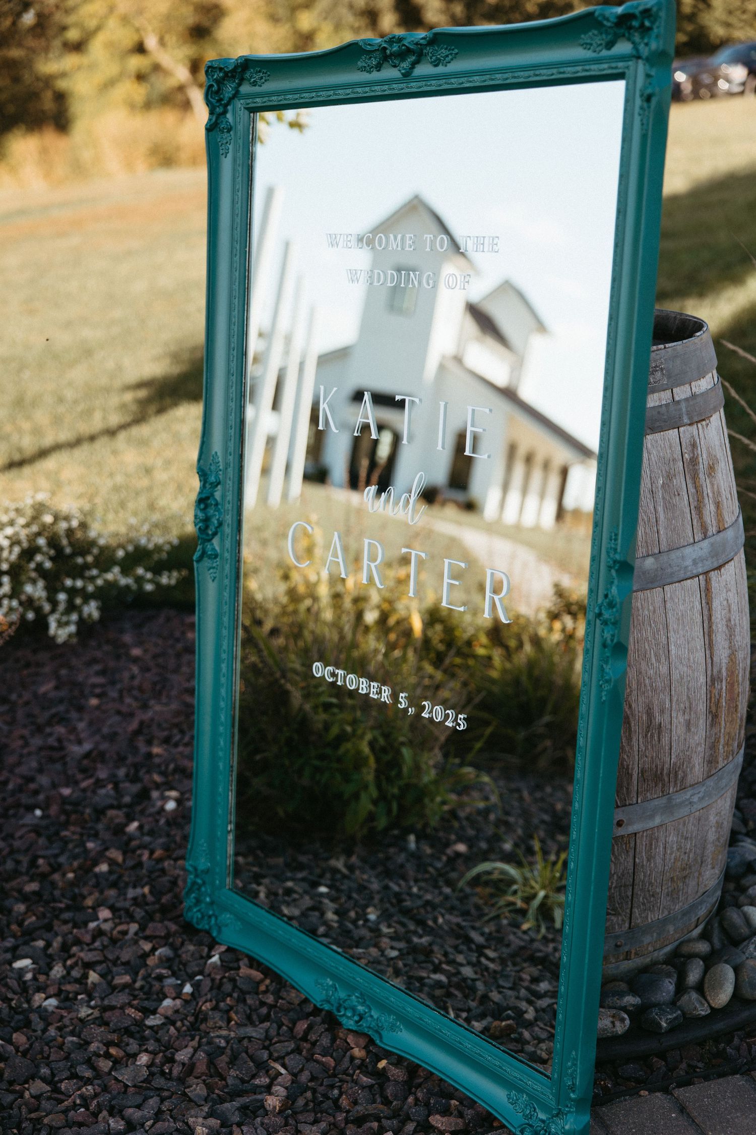 A teal-framed wedding welcome sign displayed next to a rustic wooden barrel in an outdoor setting.