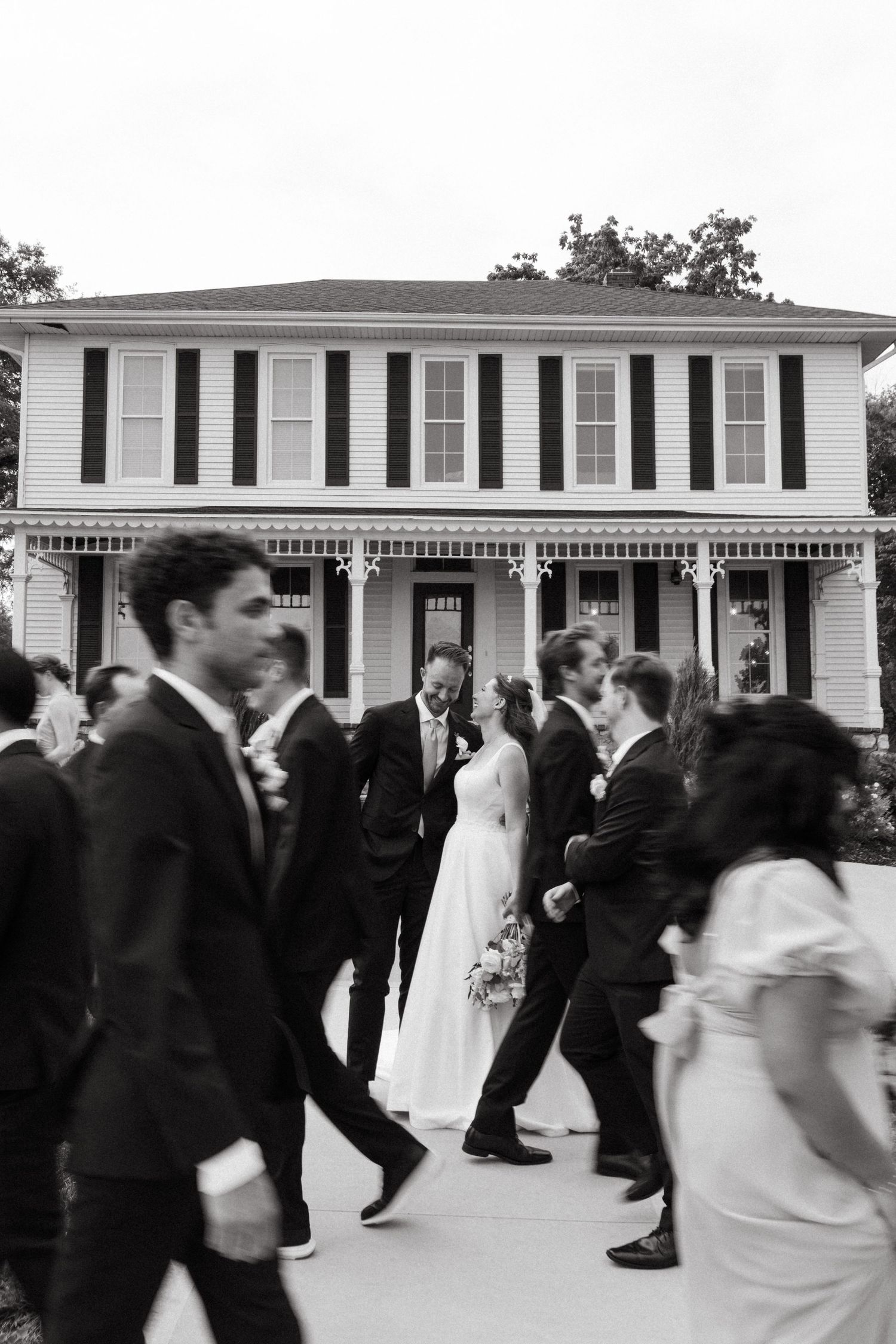 Black and white photo of wedding party walking past a historic white colonial venue after ceremony.