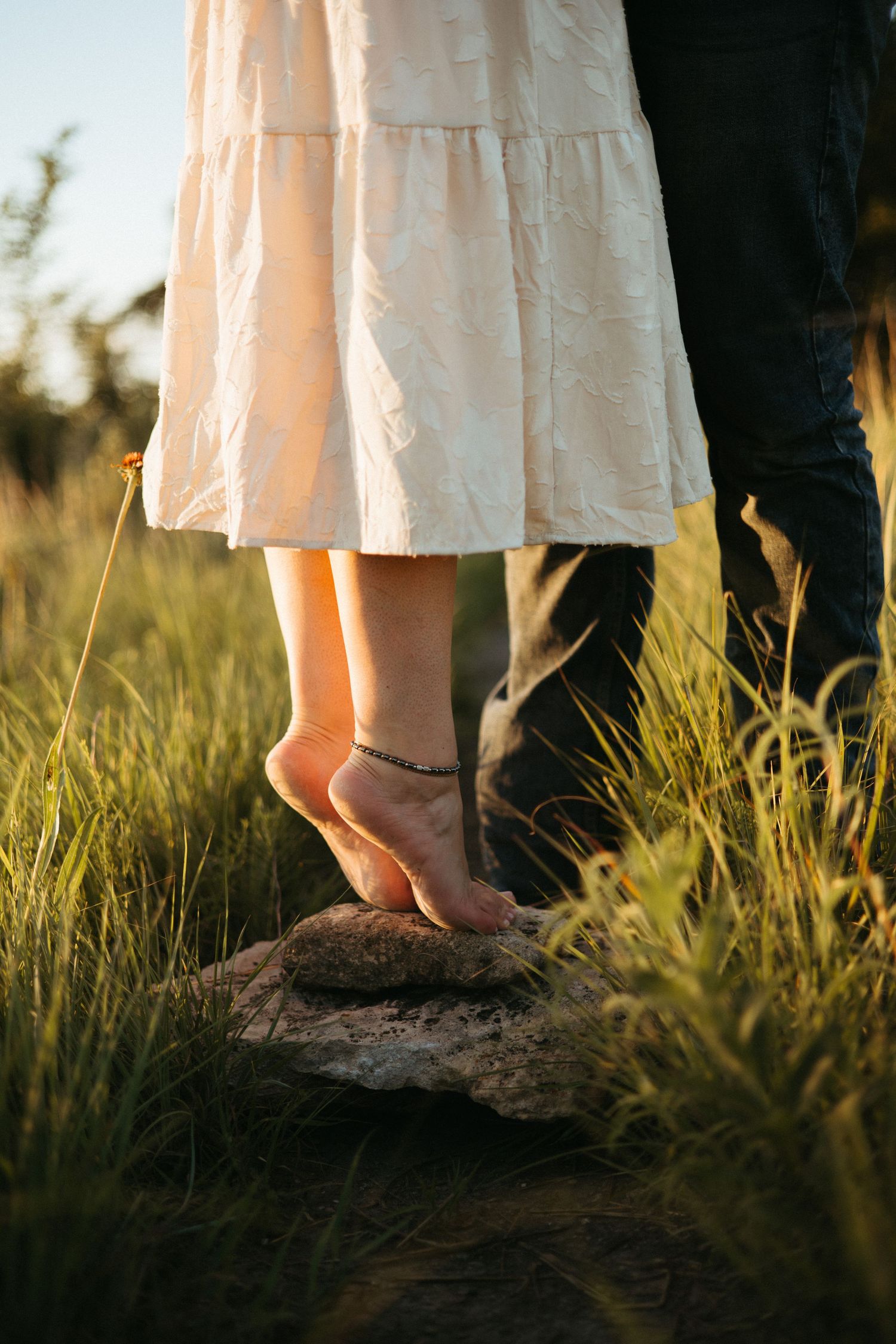 Close-up of feet in grass during sunset showing a white dress and dark pants.