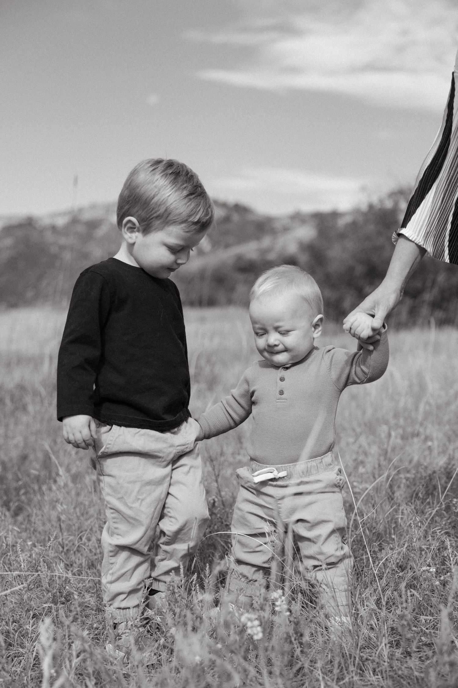 Two toddlers in light colored clothing play together in a grassy meadow with hills in the background.