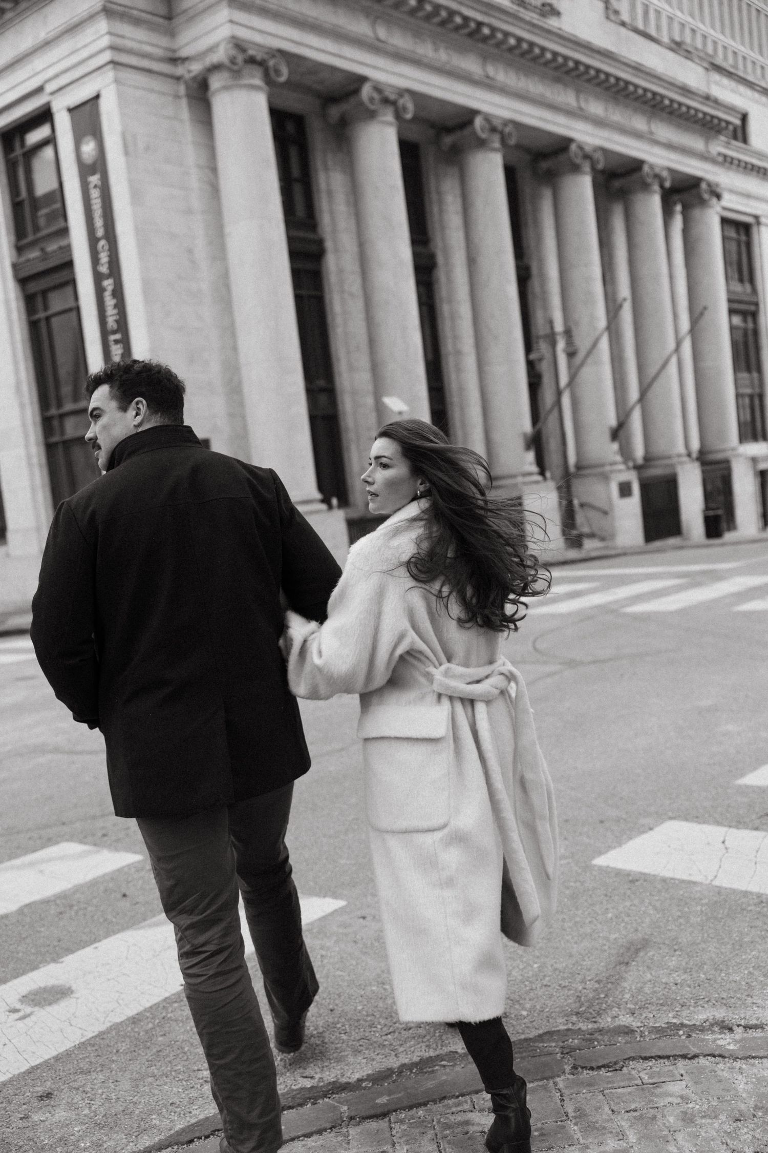 A couple walking past a neoclassical building with columns, with wind blowing through her hair in black and white.