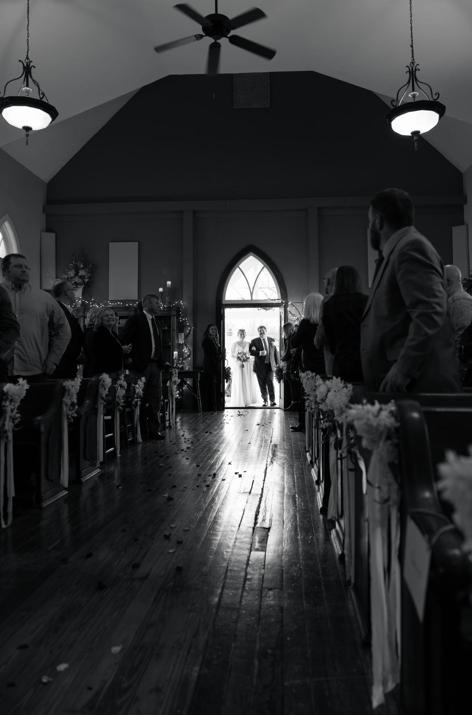 Black and white photo of a wedding ceremony in a chapel with silhouettes in the arched doorway and wooden floors.