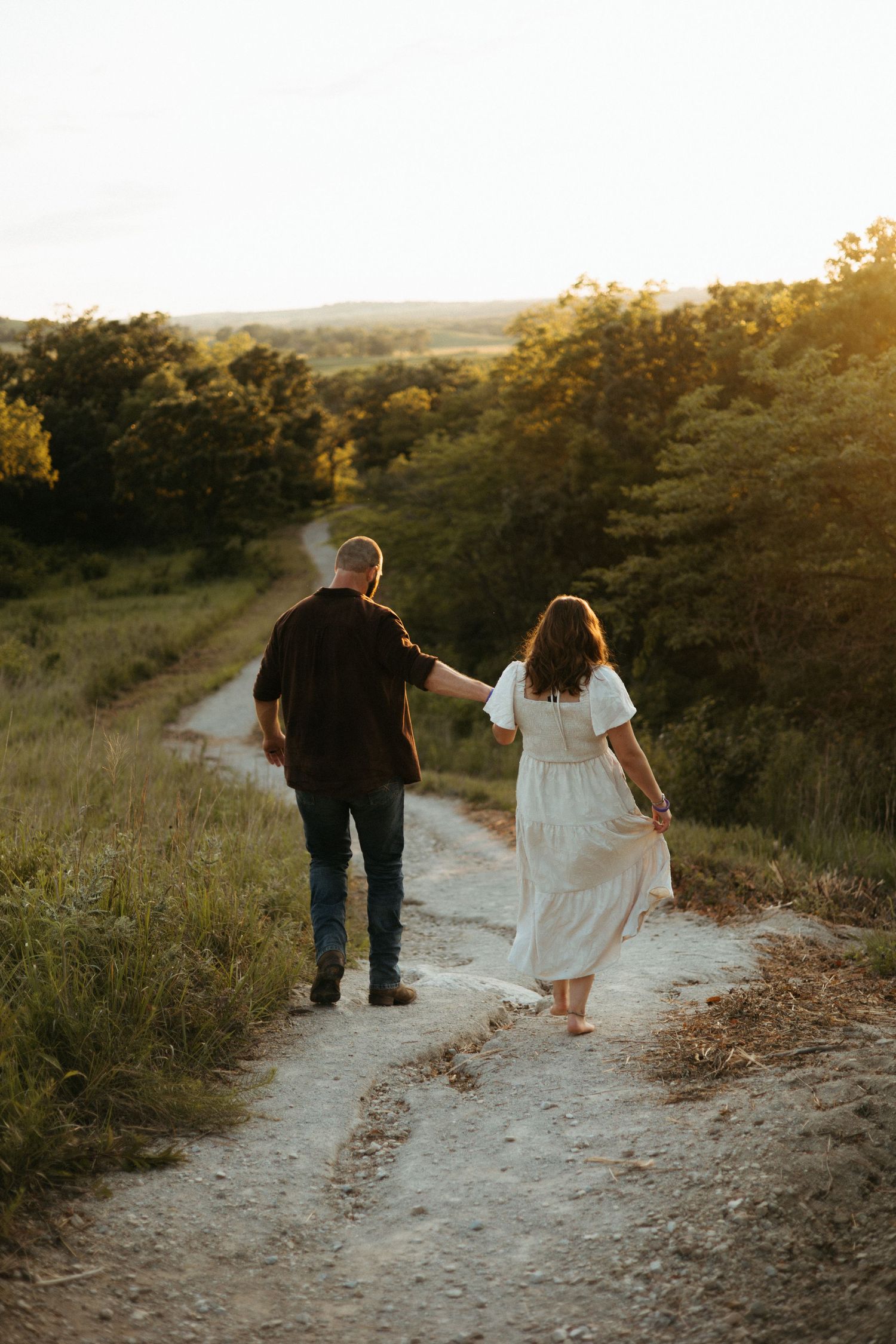 A couple walks hand in hand along a winding dirt path during golden hour in a scenic natural setting.