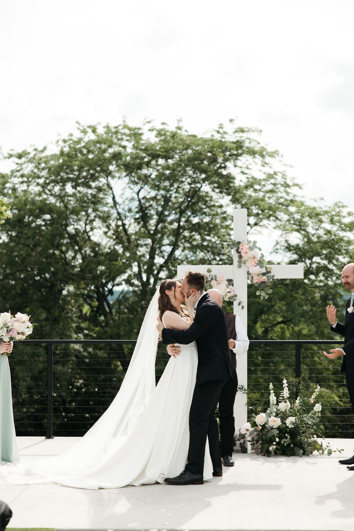 Bride and groom share first kiss at outdoor wedding ceremony with white cross backdrop and lush green trees.