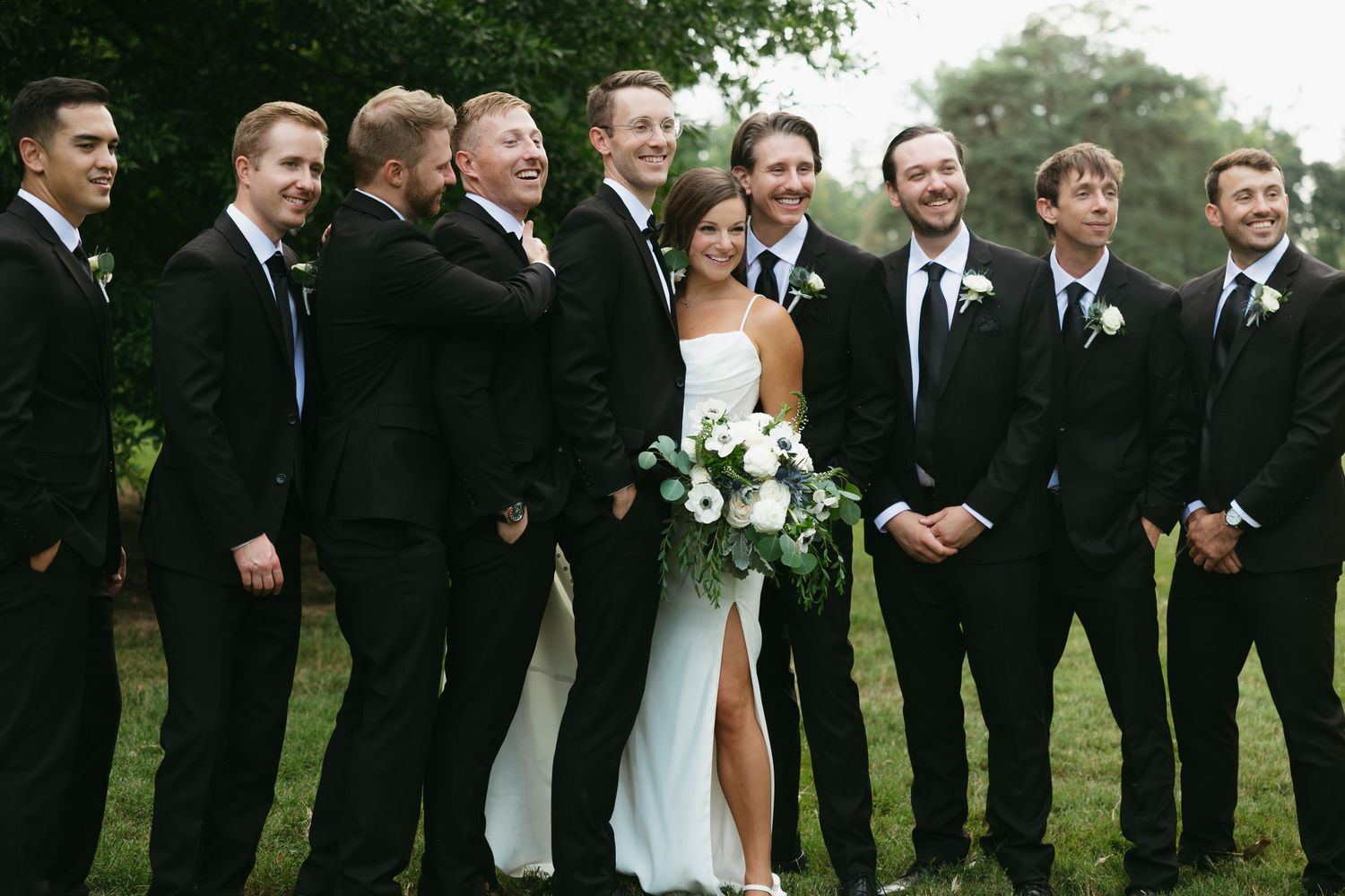 Wedding party in formal black and white attire stands together outdoors in a line against green foliage.
