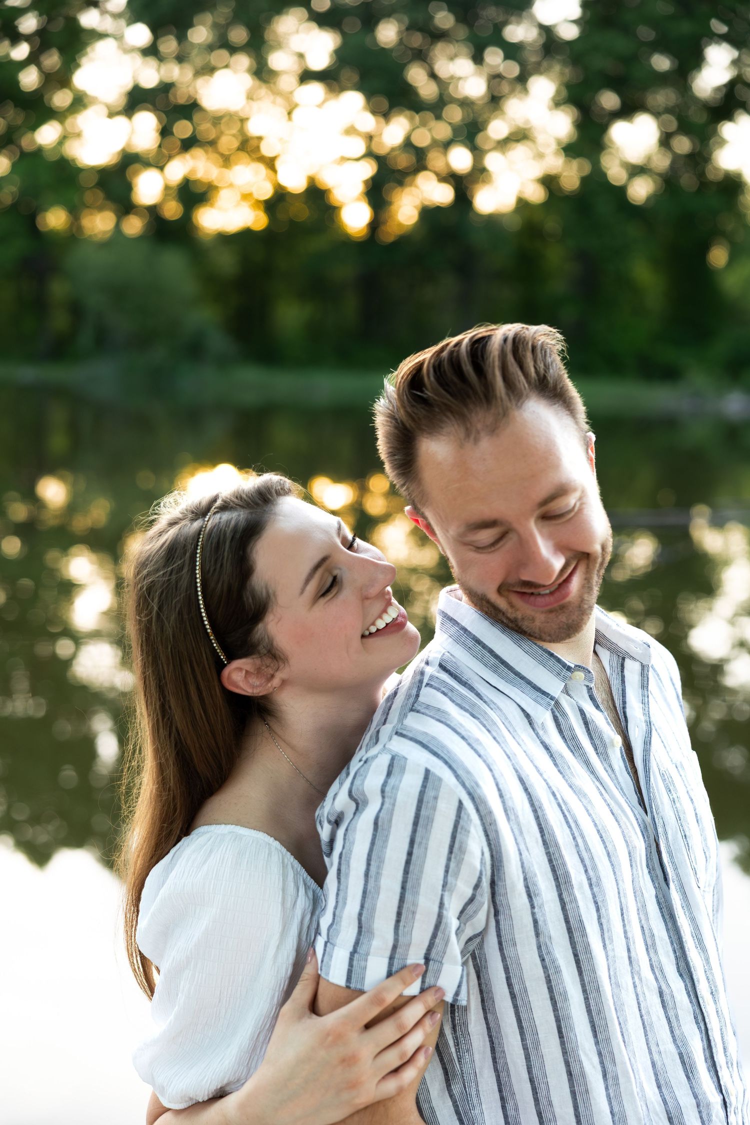 A couple shares tender moments by a lake during golden hour, wearing white and striped clothing.