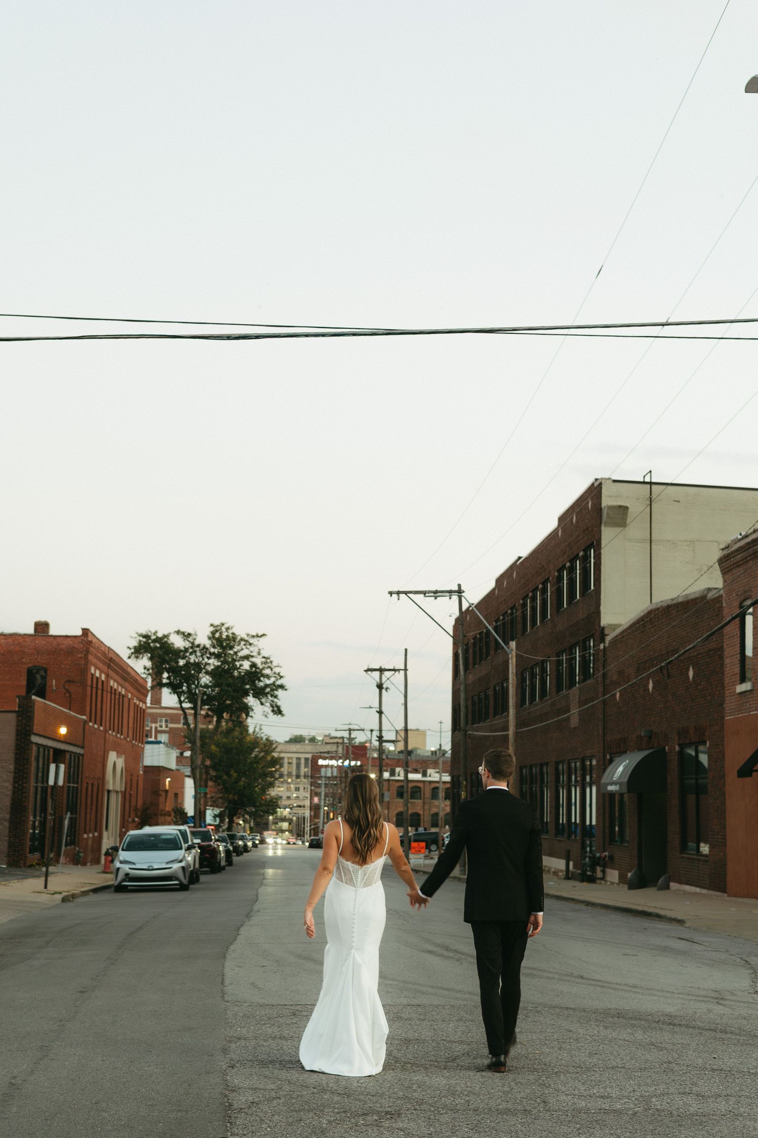 A couple in wedding attire walking down an urban street lined with brick buildings and power lines.