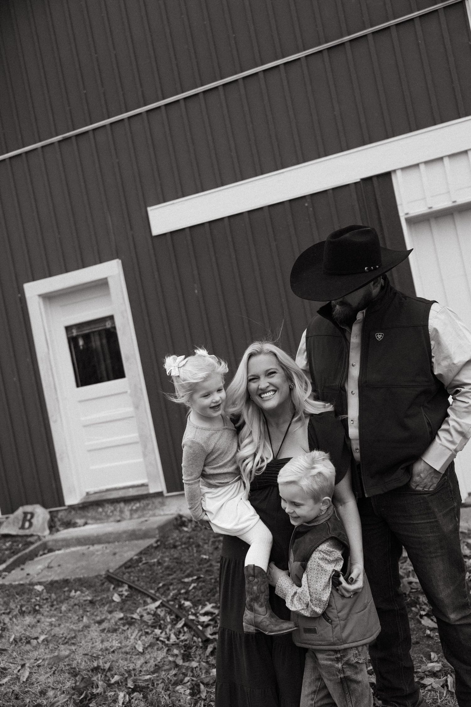 A family stands together in front of a rustic barn with dark siding and white trim in a black and white photograph.