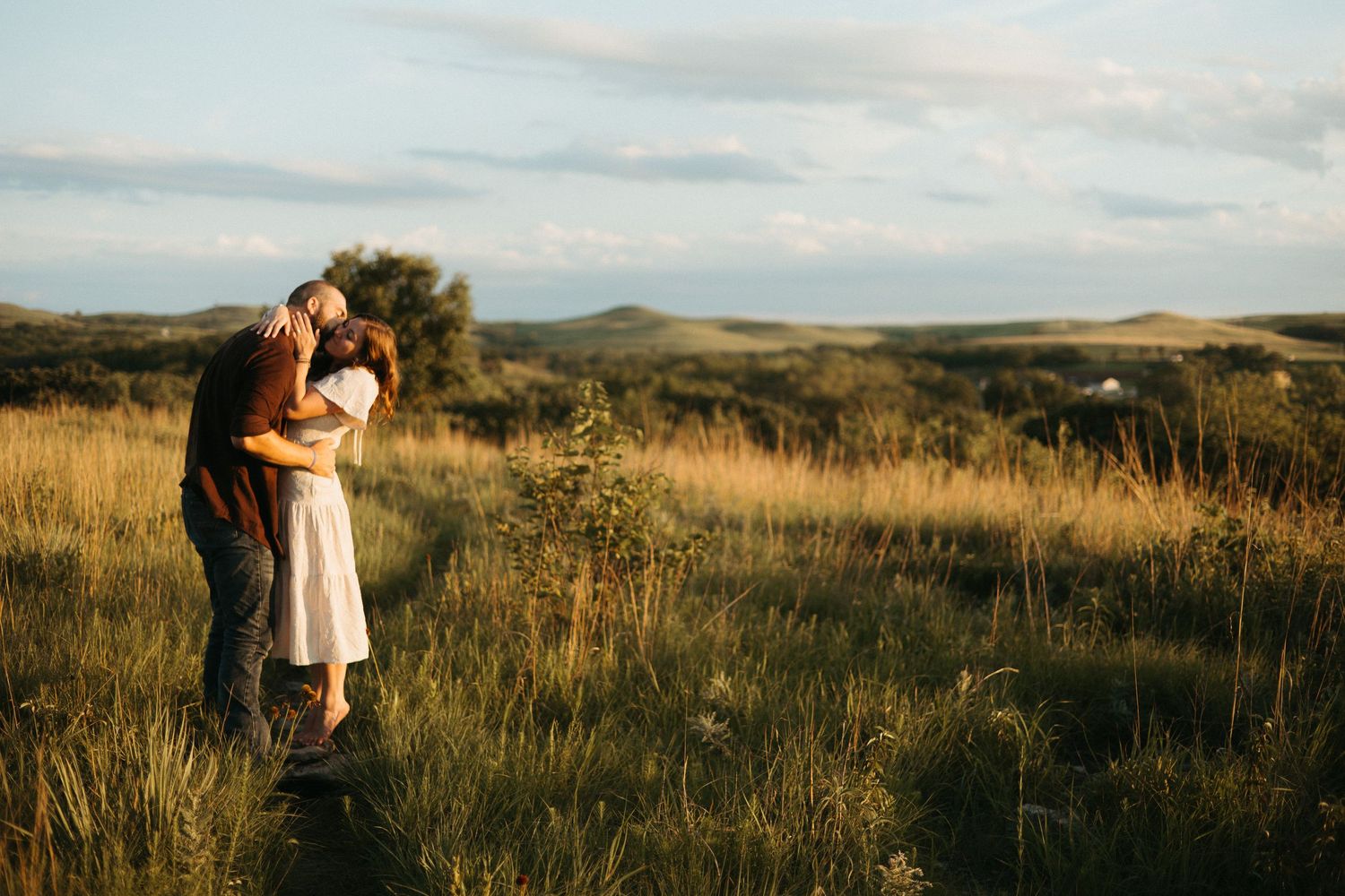 A couple embraces in a golden field at sunset with mountains in the background.