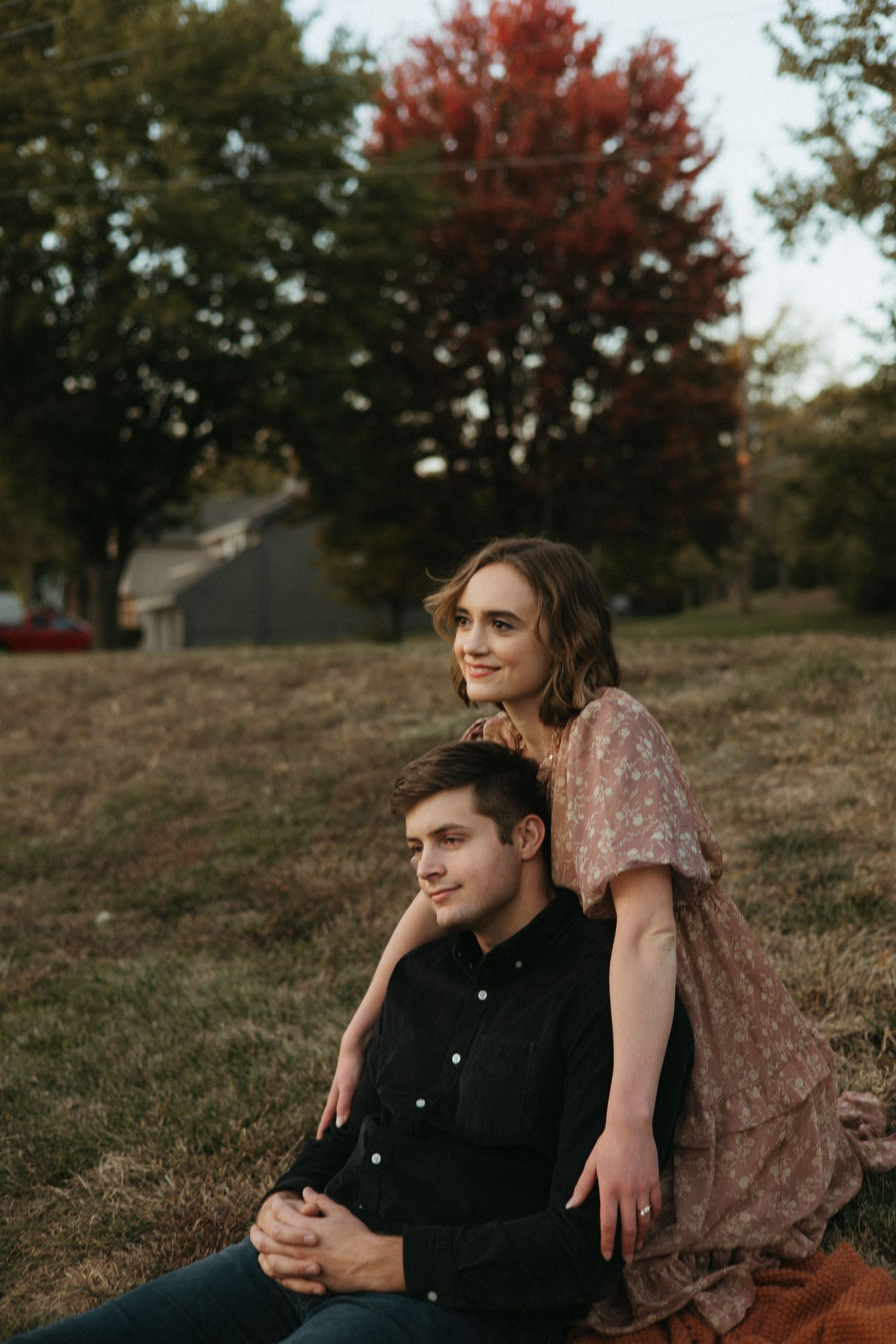A couple sits together on a grassy hillside during autumn with red maple trees in the background.