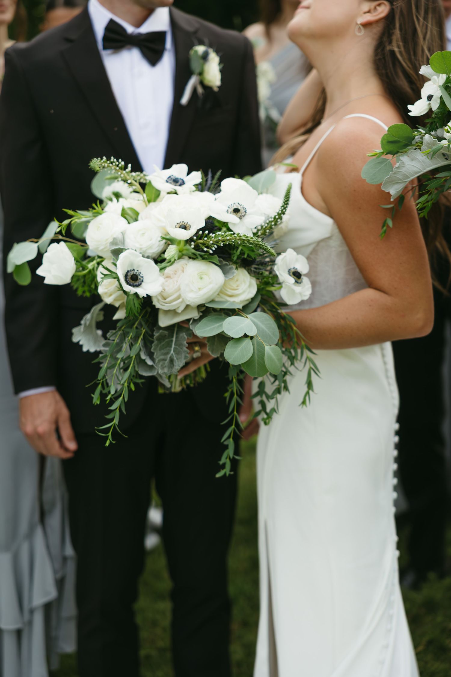 Wedding party members in formal black tuxedos and white dresses hold elegant white and green floral bouquets outdoors.