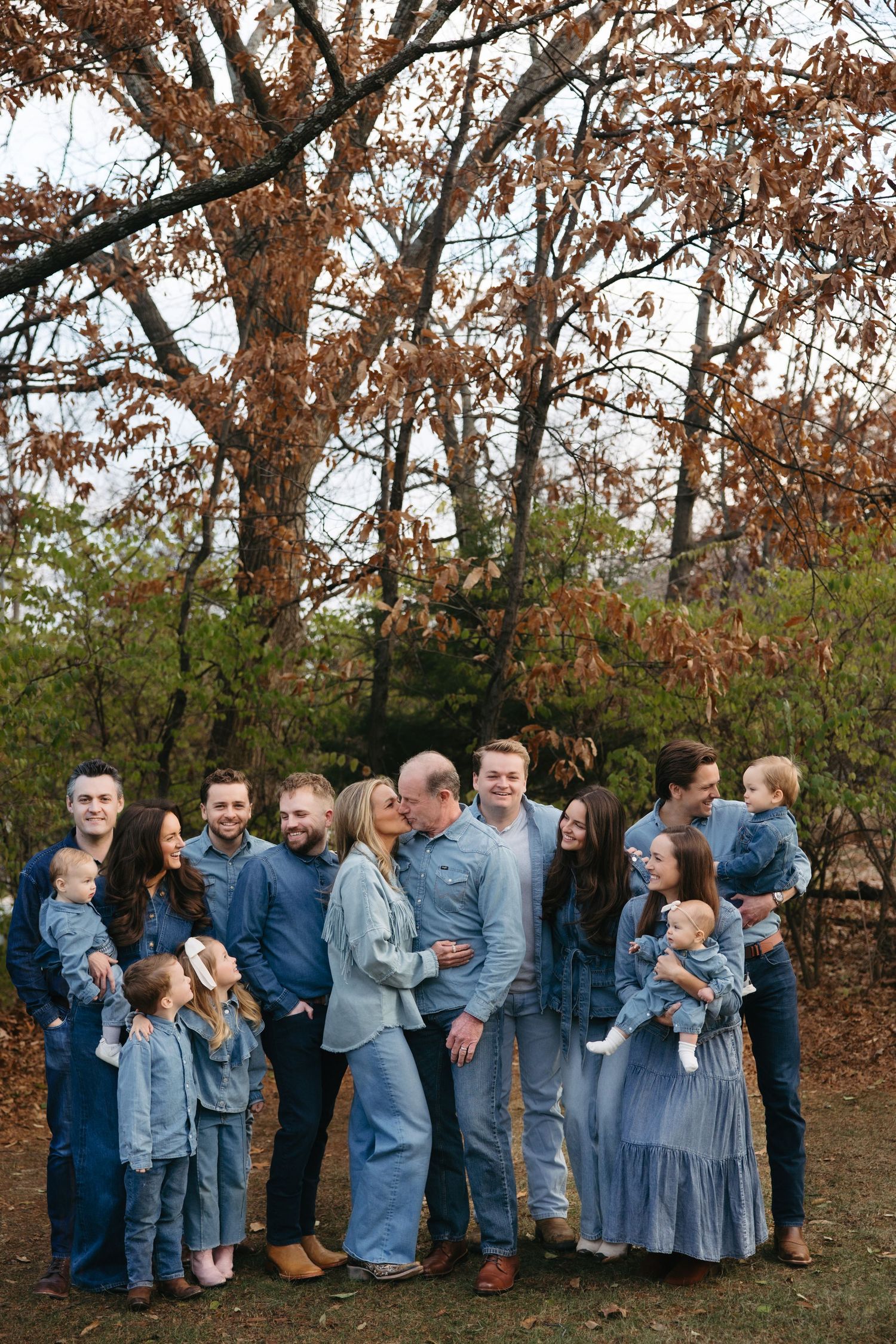 Large family posing outdoors in coordinating denim outfits during autumn, surrounded by trees with fall foliage.