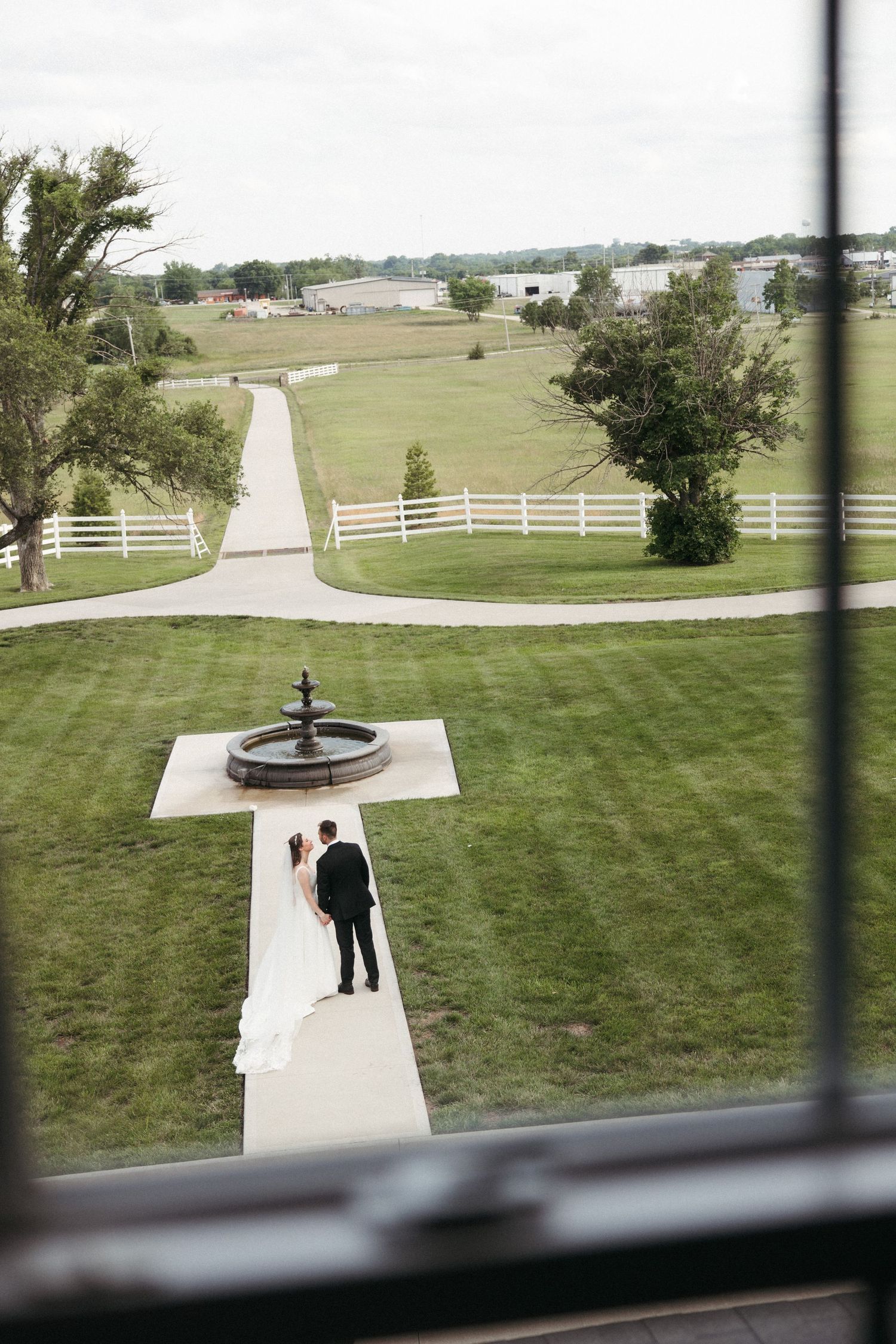 Aerial view of bride and groom standing near a fountain on a white pathway surrounded by lush green fields and white fencing.