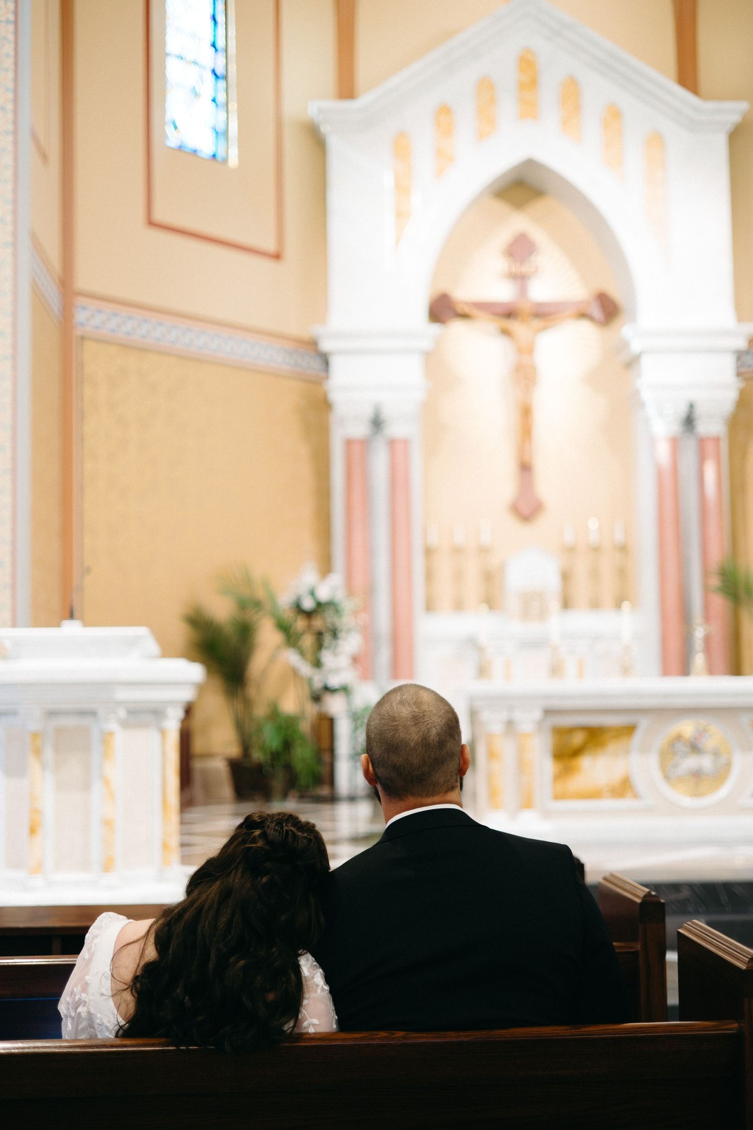People sit in church pews during a service in a traditional Catholic church with ornate architecture and altar.