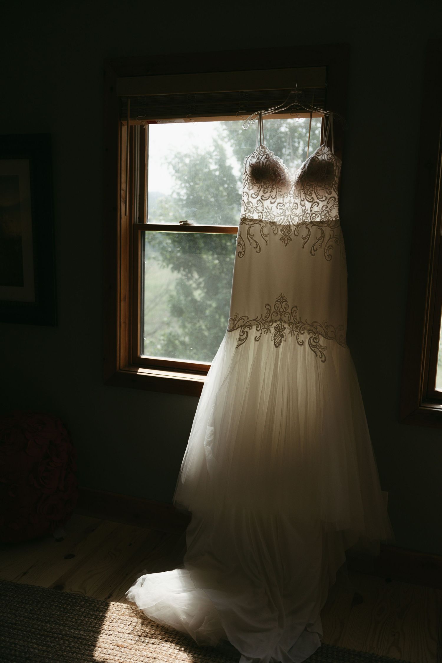 A wedding dress hanging in silhouette against natural window light in a dark room.