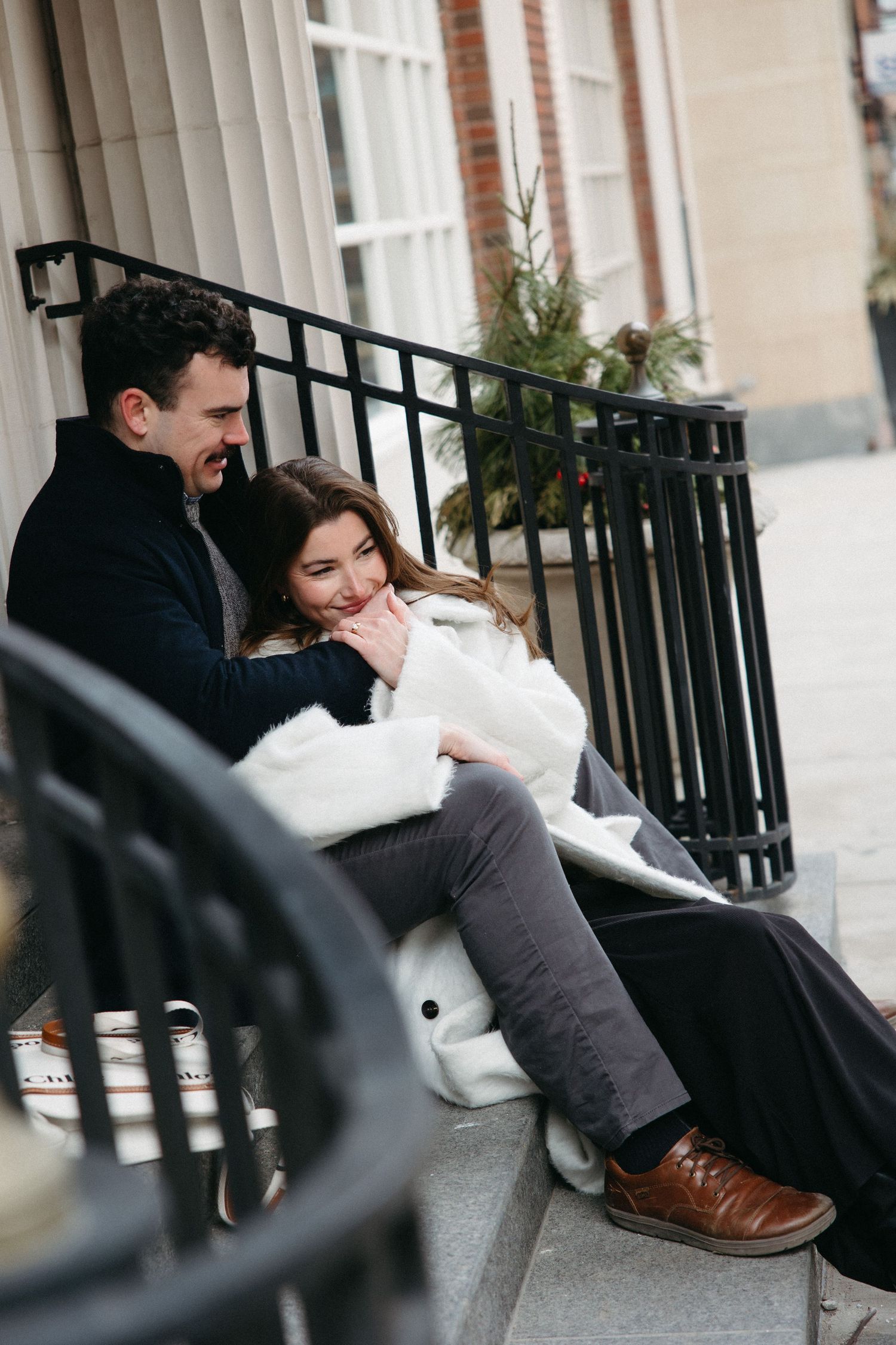 An intimate couple resting together on concrete steps near an iron railing and white columns.