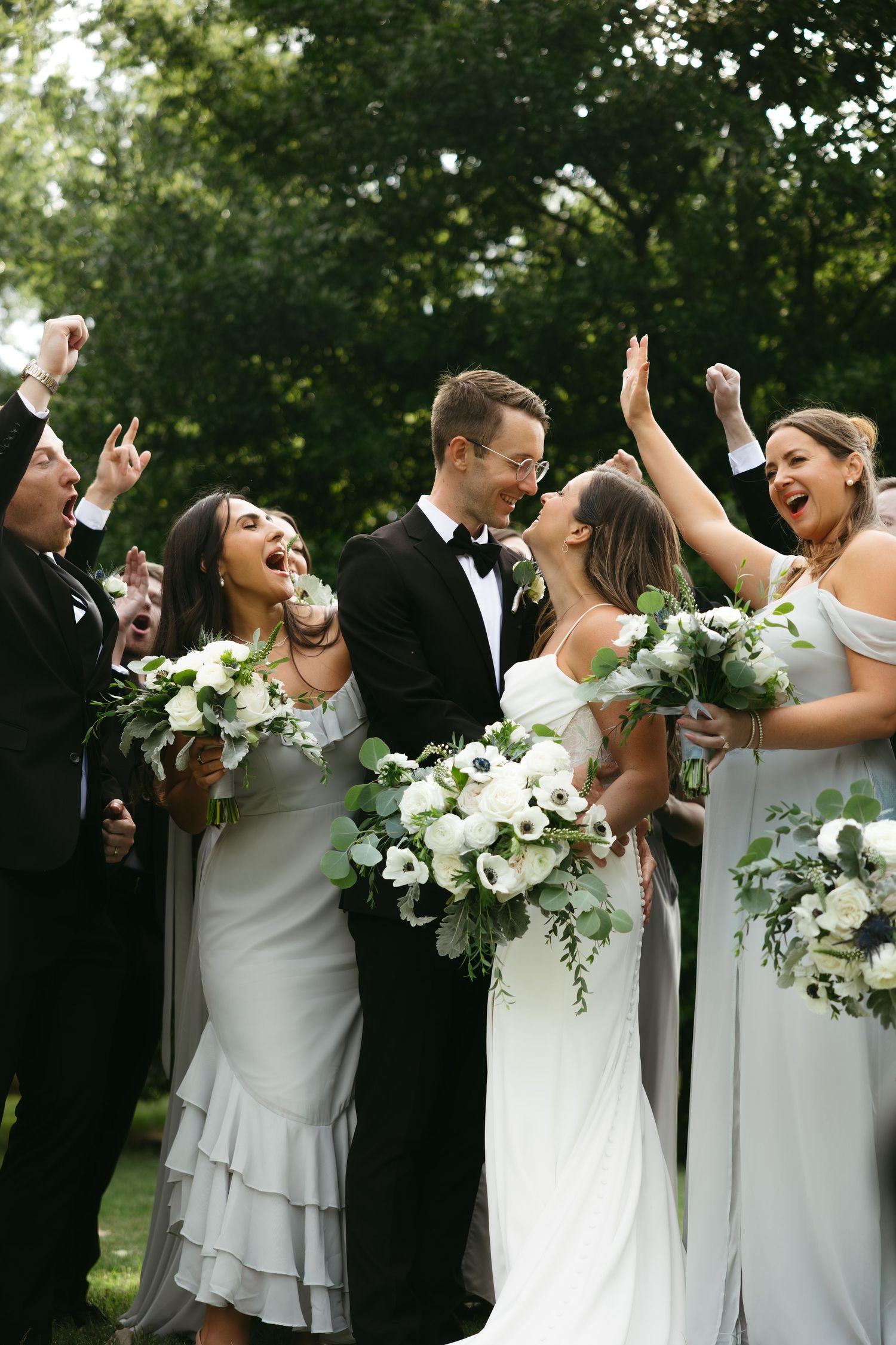 Wedding party in gray dresses and black tuxedos laughing and celebrating together outdoors with white floral bouquets.