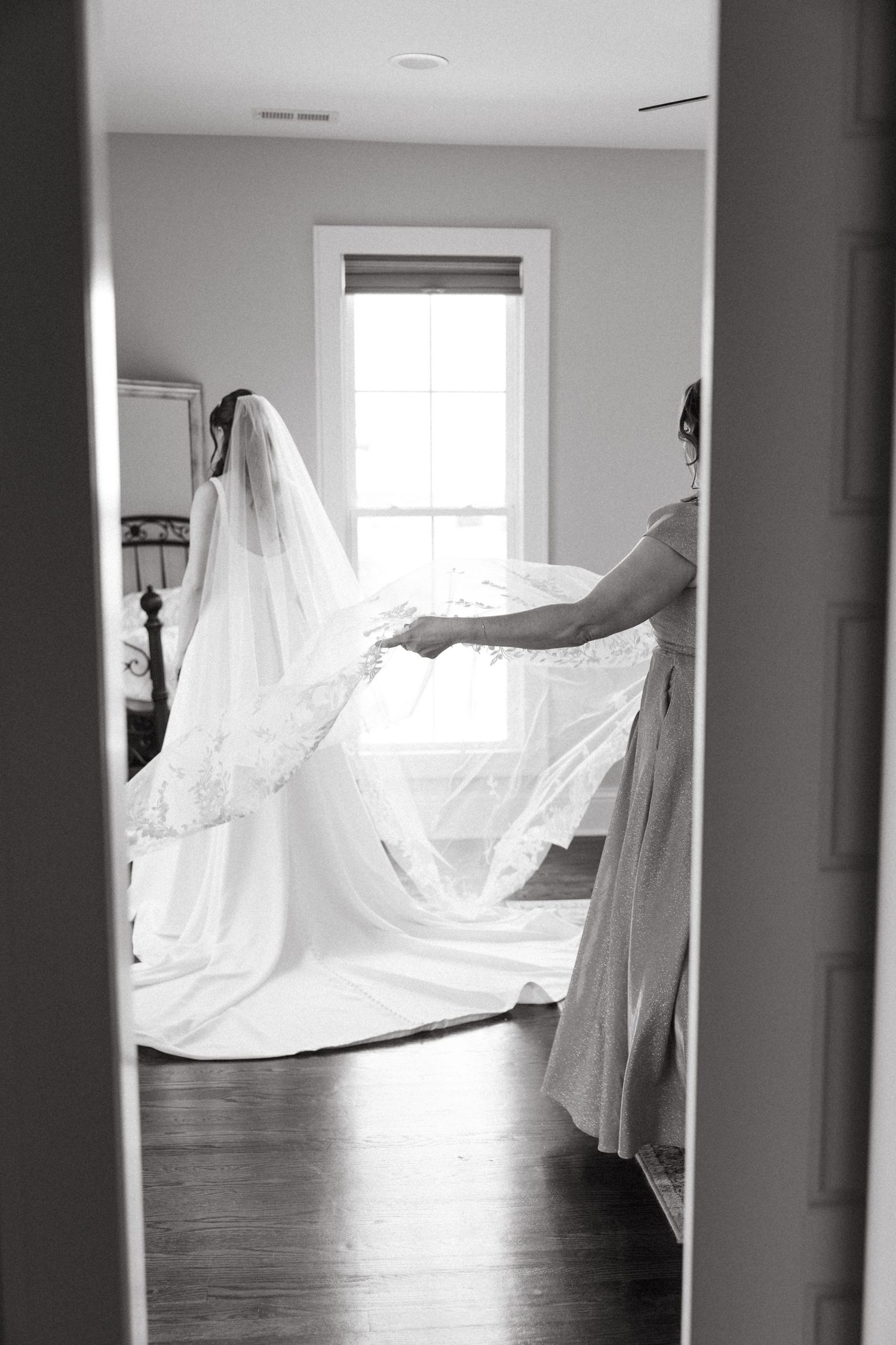 Black and white photo of a bride in a flowing gown and veil being helped by a bridesmaid in a bright white room.