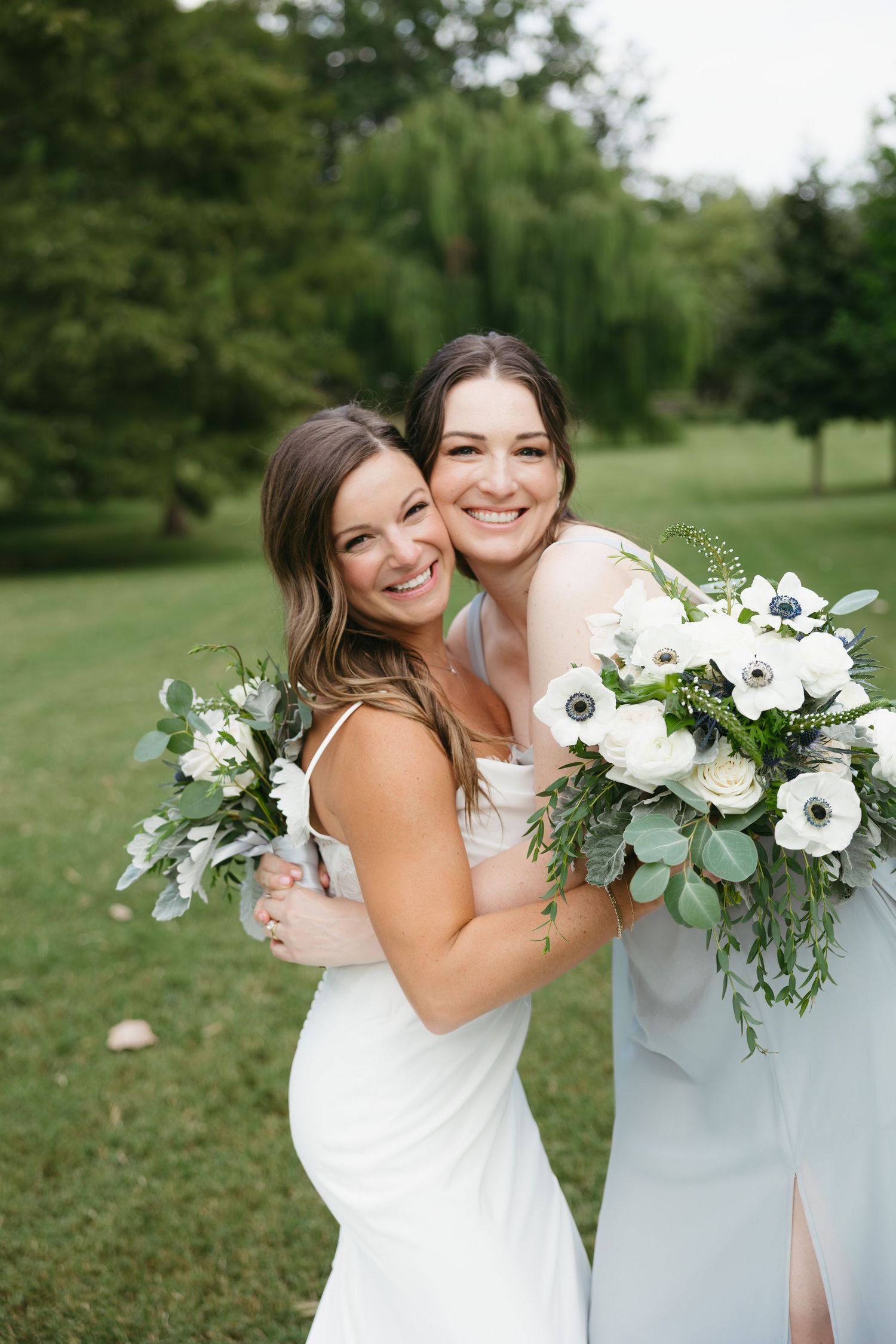 Two people in light-colored dresses embrace while holding white floral bouquets in an outdoor garden setting.