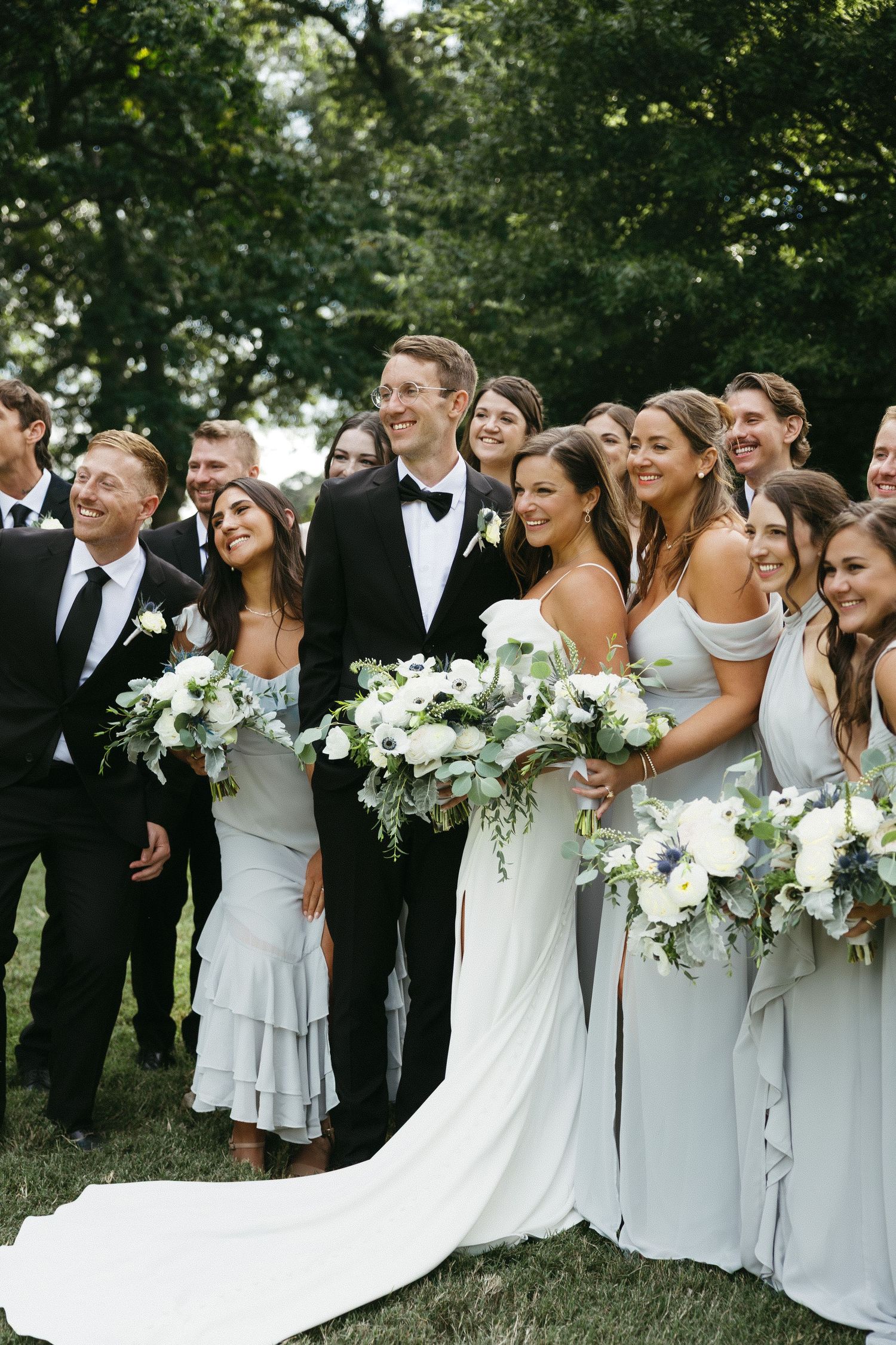 Wedding party in light blue dresses and black tuxedos pose together outdoors holding white floral bouquets.