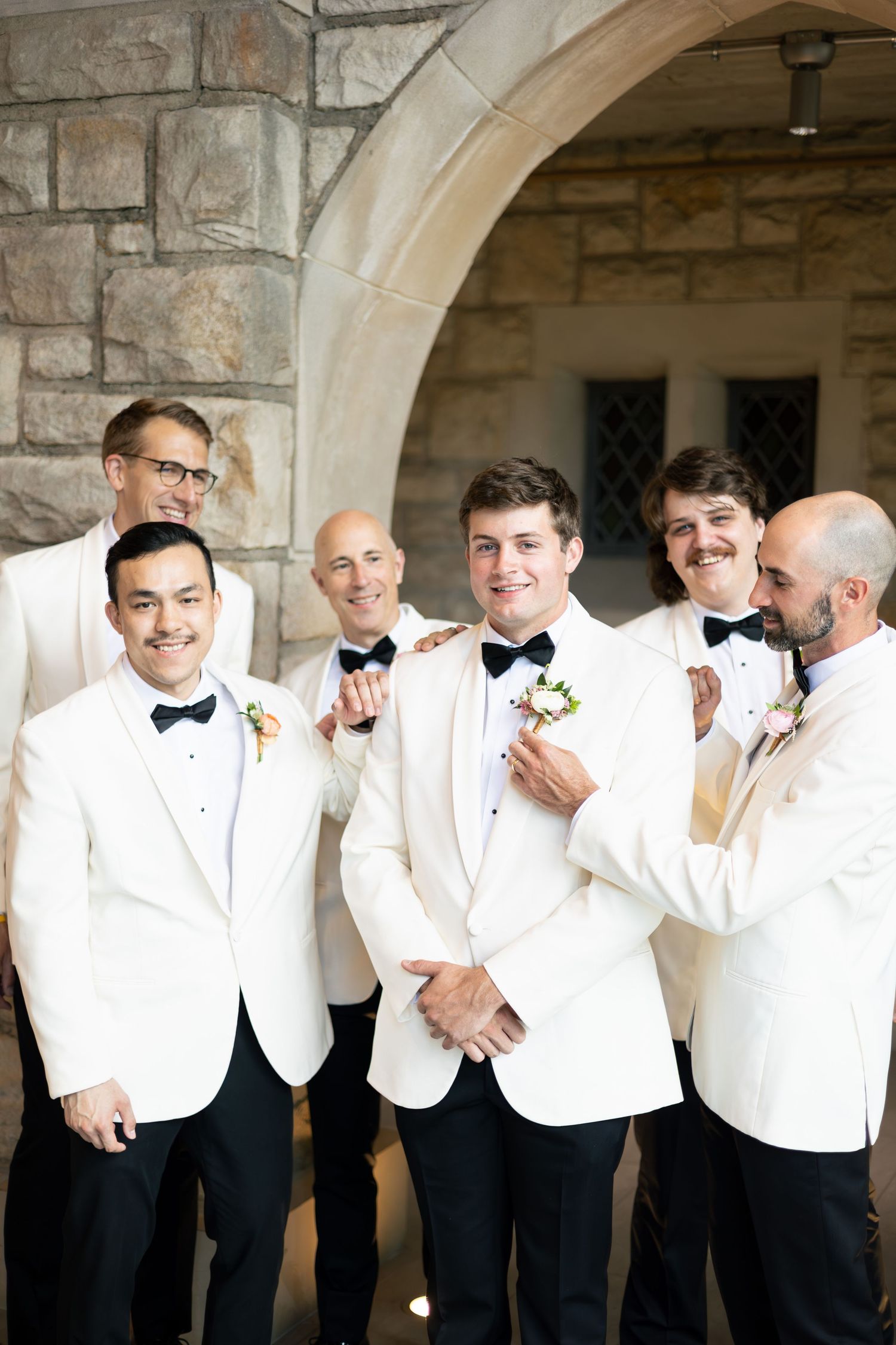 Groomsmen in white dinner jackets and black bow ties pose under stone archways.