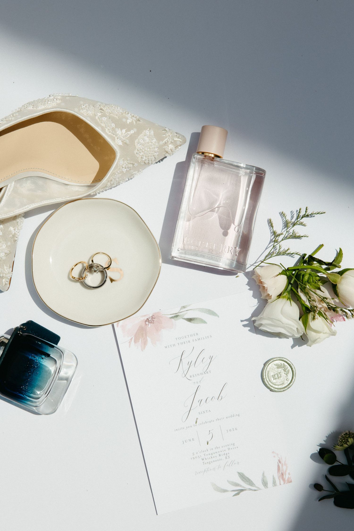 Flat lay of wedding details including rings, perfume, invitation, and white flowers.