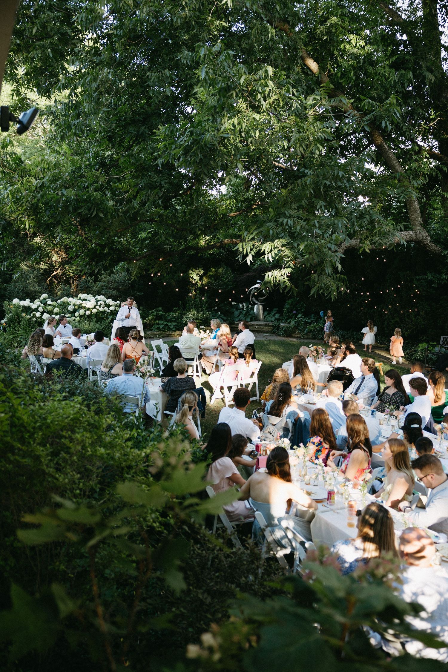 A large group gathers for an elegant outdoor dinner party under tall trees at dusk.