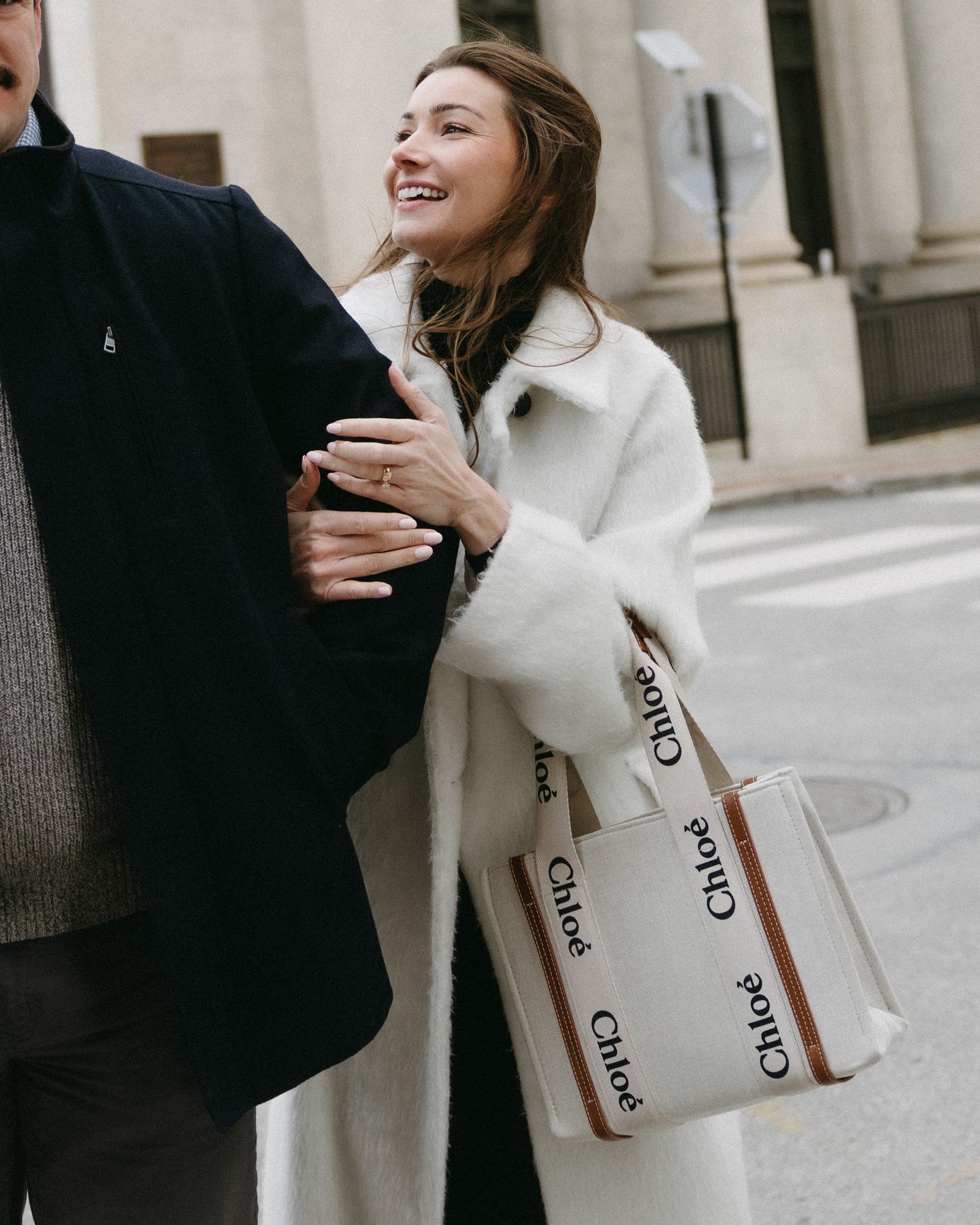 A person in a white winter coat laughs while holding a Chloe shopping bag on a city street.