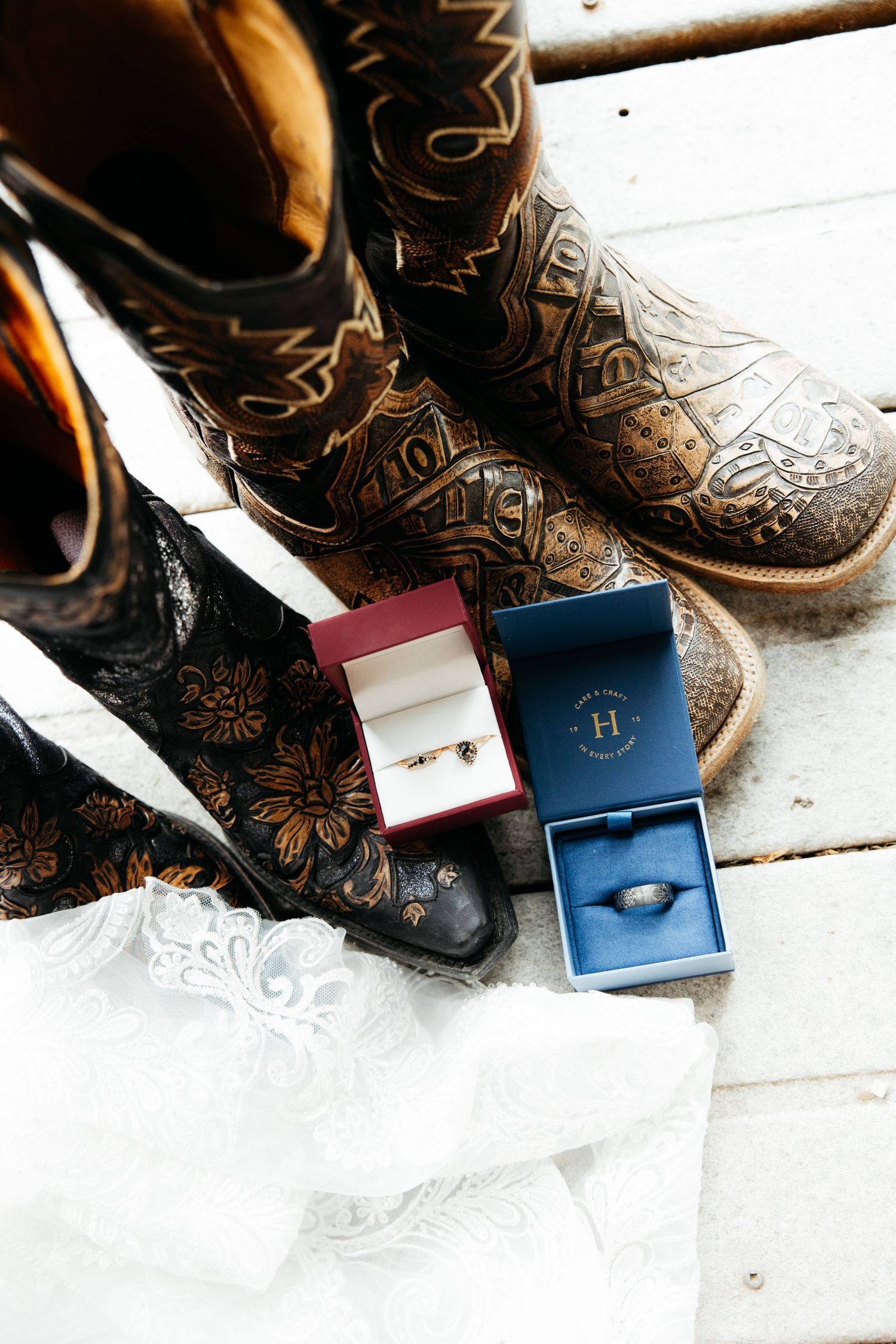 Wedding rings in blue boxes placed next to decorative cowboy boots on a white surface.