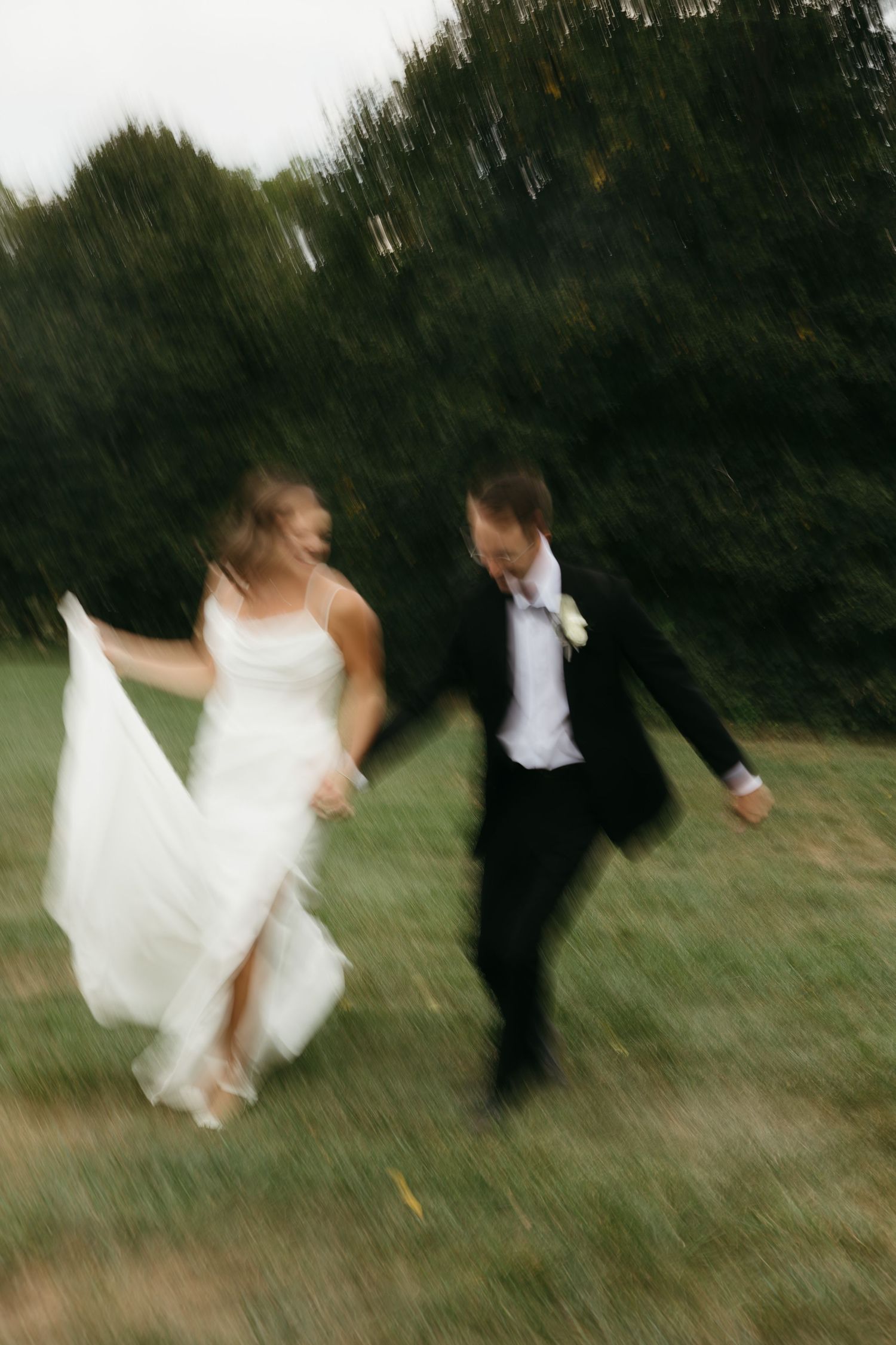 A blurred motion shot of newlyweds running and dancing together on grass with trees in the background.