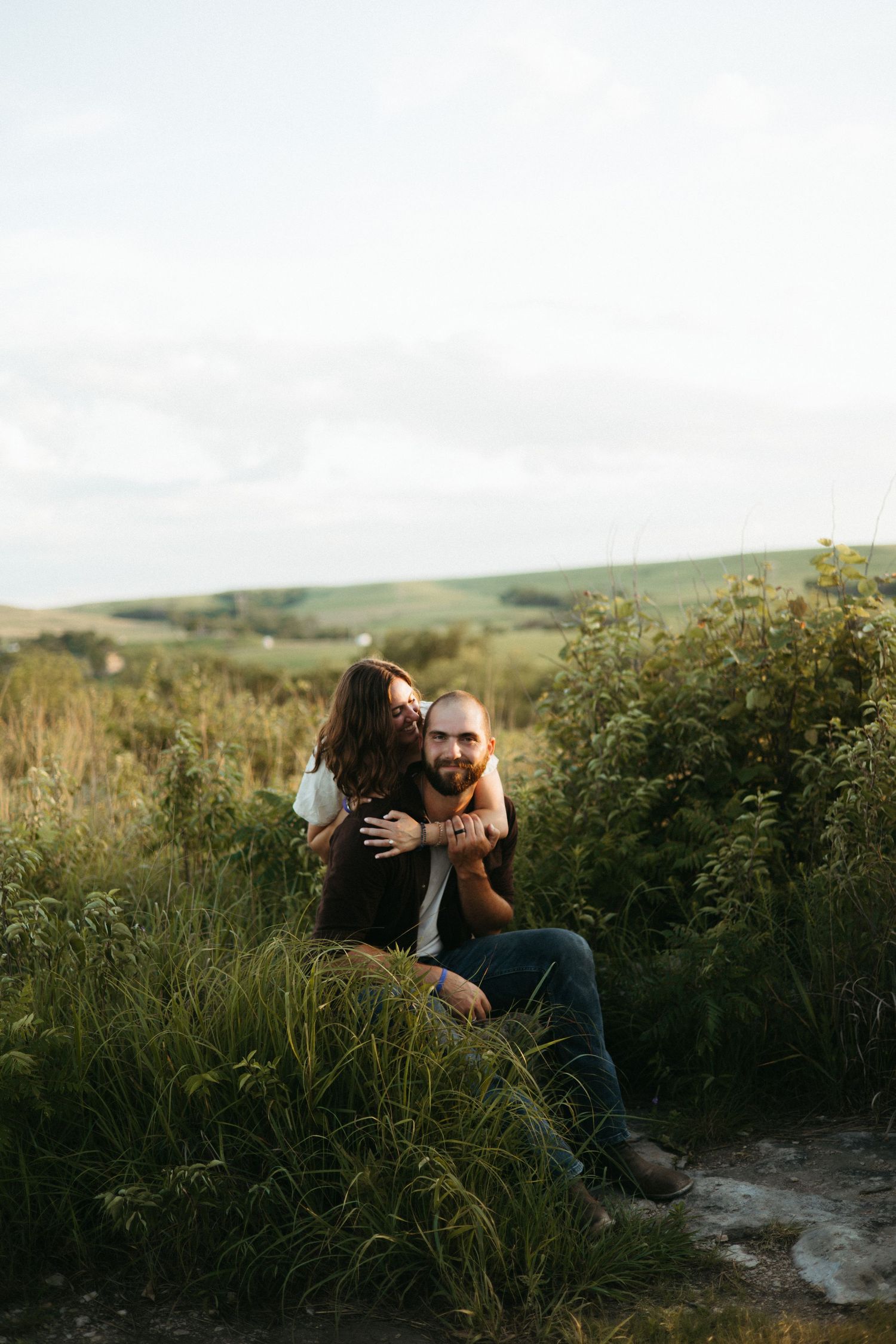 A romantic couple embraces against a scenic natural backdrop of rolling hills and greenery at golden hour.
