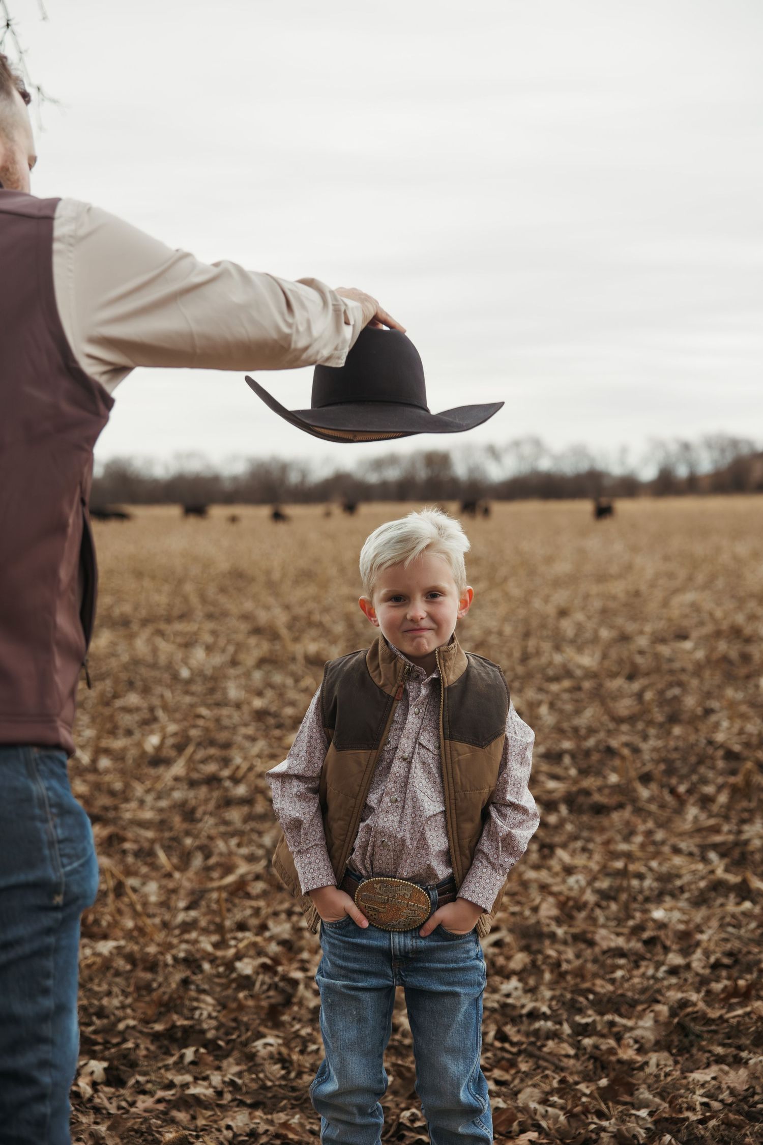 A young child wearing a cowboy hat and vest stands in a plowed field during autumn.