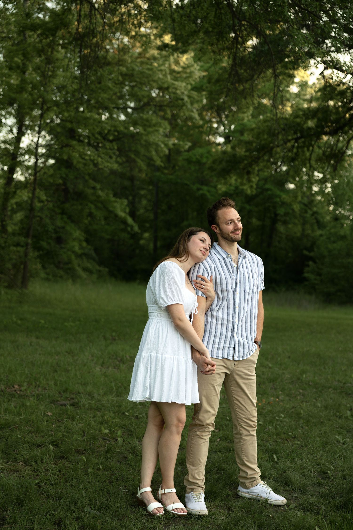 A couple stands together in a lush green park setting wearing coordinating white and striped outfits.